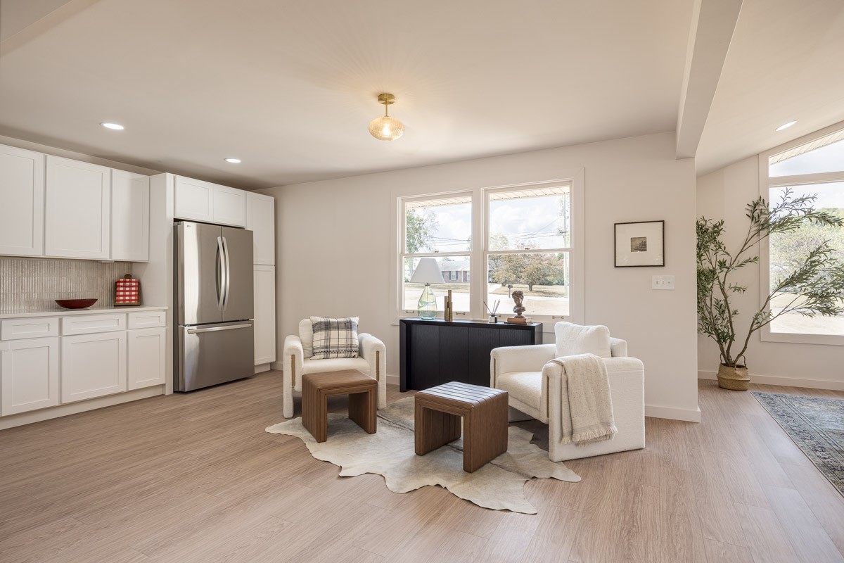 Open-plan kitchen and sitting area with white cabinets, stainless steel refrigerator, and two armchairs by large windows.