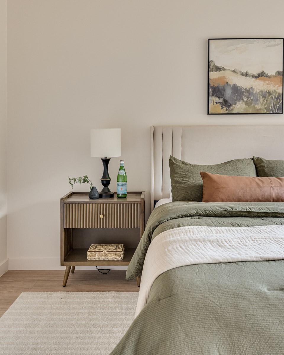 Bedroom with muted green bedding, an upholstered headboard, and a wood nightstand beneath abstract art.