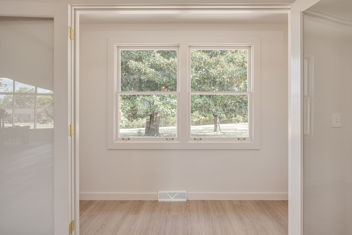 Empty room with light wood flooring, two windows overlooking leafy trees, and a floor vent.