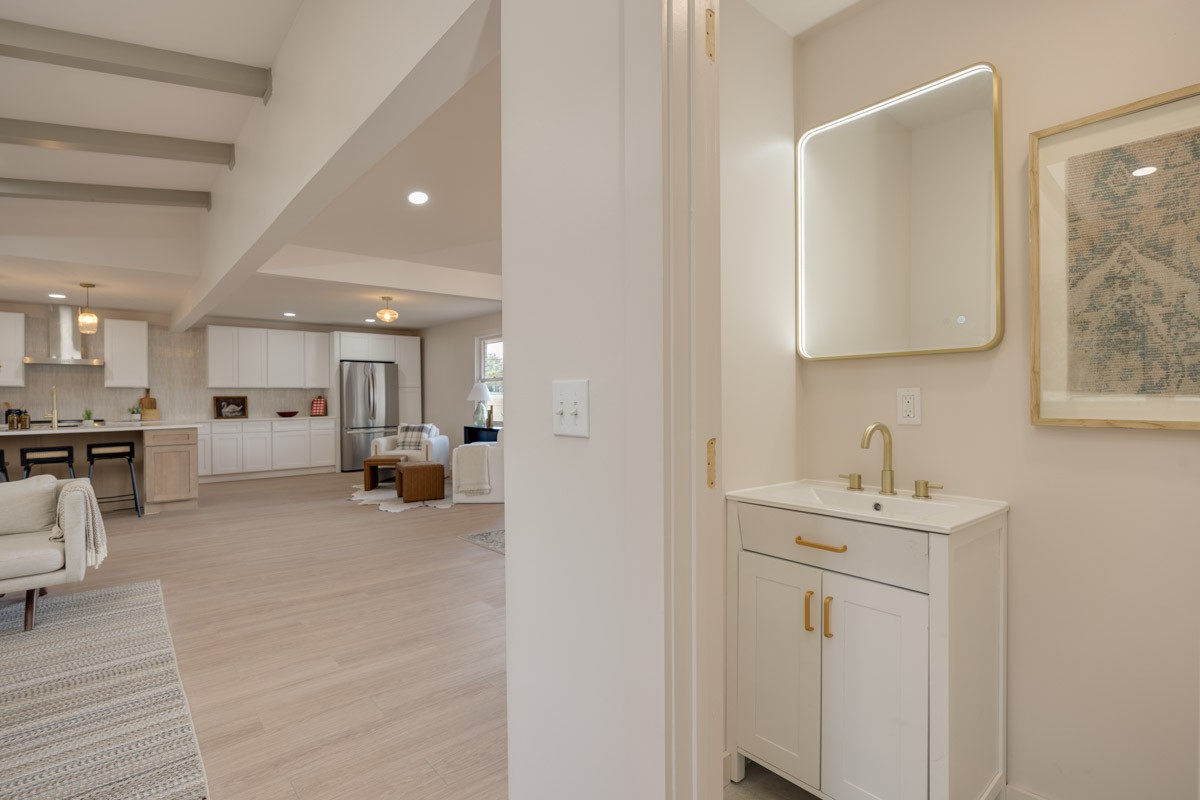 Bathroom vanity with brass fixtures and illuminated mirror overlooking an open kitchen and living area.