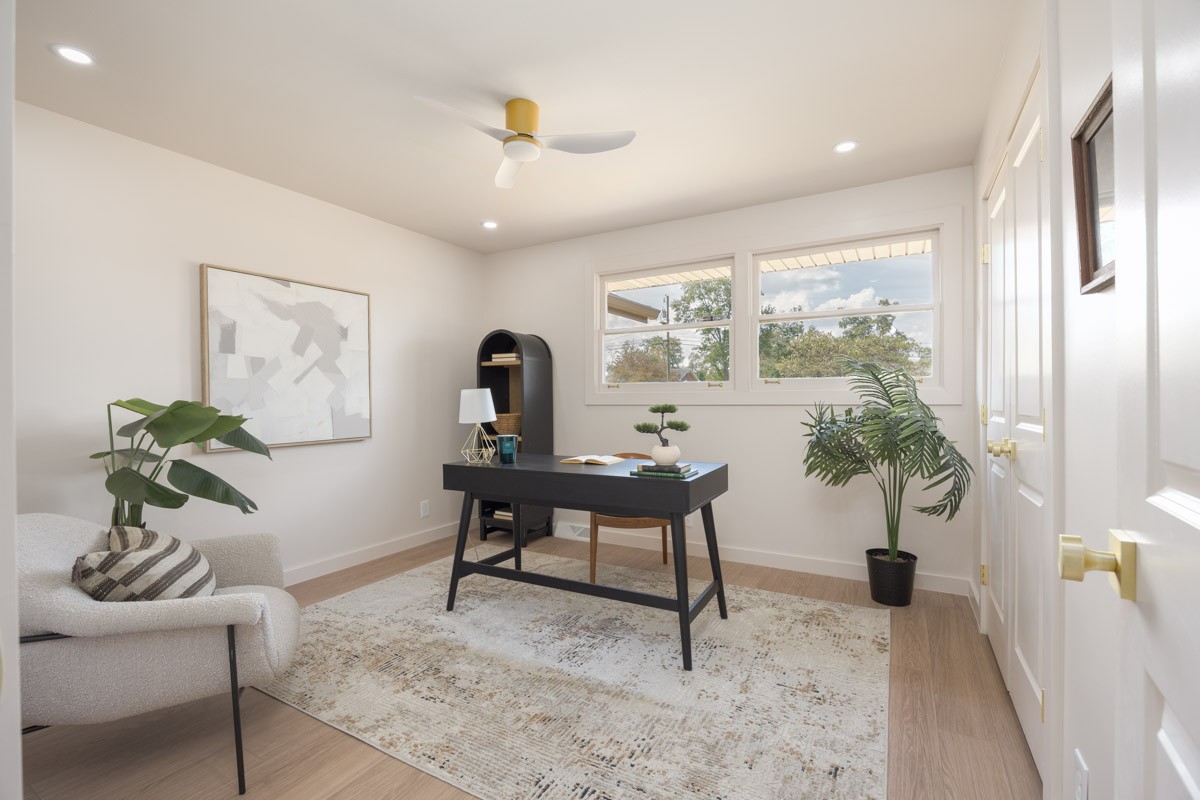 Staged home office with a black desk, white armchair, potted plants, and two windows.