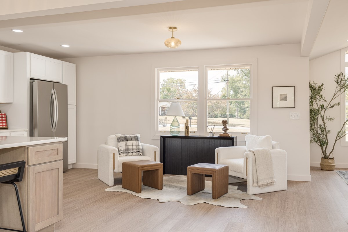 Staged living room with two white armchairs, wooden tables, and a black console beneath double windows.