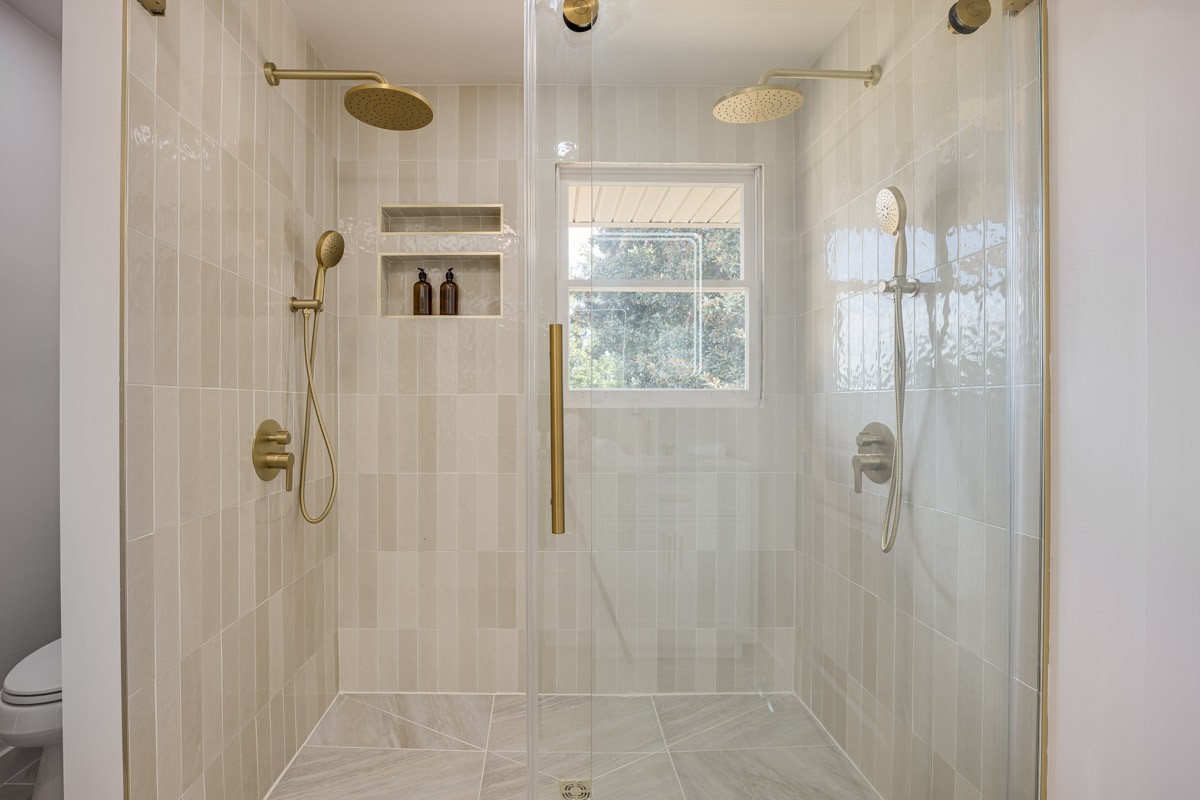 Walk-in tiled shower with glass enclosure, brass fixtures, built-in niches, and a window.