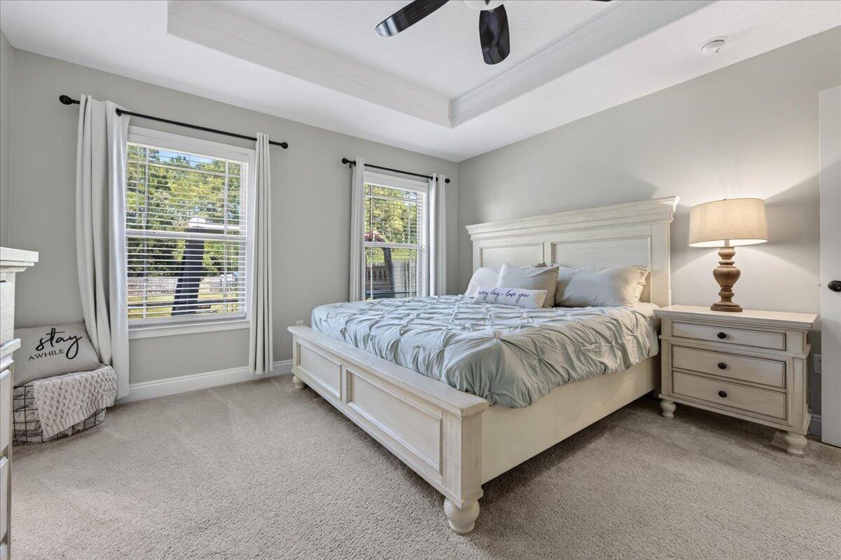 Staged bedroom with a white paneled bed, pale blue-gray bedding, and two windows overlooking greenery.