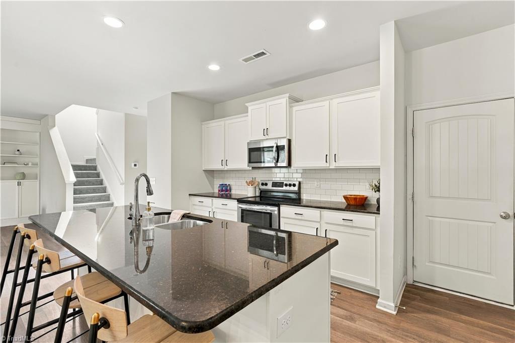 Kitchen with white shaker cabinets, dark stone island, stainless steel appliances, and stairs nearby.