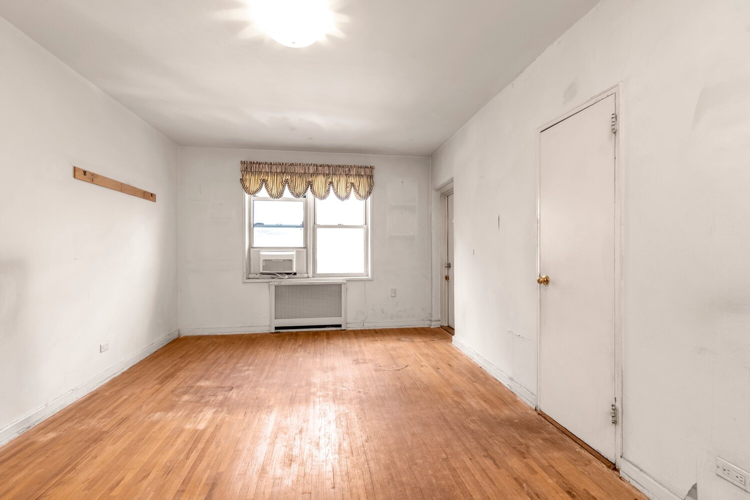 Empty apartment room with hardwood floors, a window air conditioner, and a radiator cover.