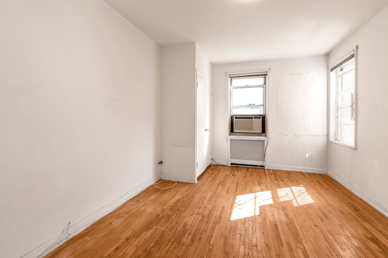 Vacant apartment room with hardwood floors, two windows, and a window air conditioner.