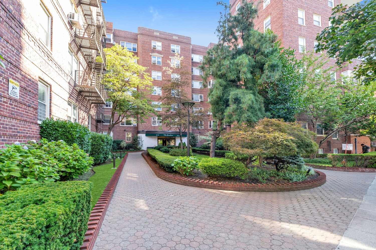 Red-brick apartment building entrance beyond a landscaped courtyard and paved walkway.