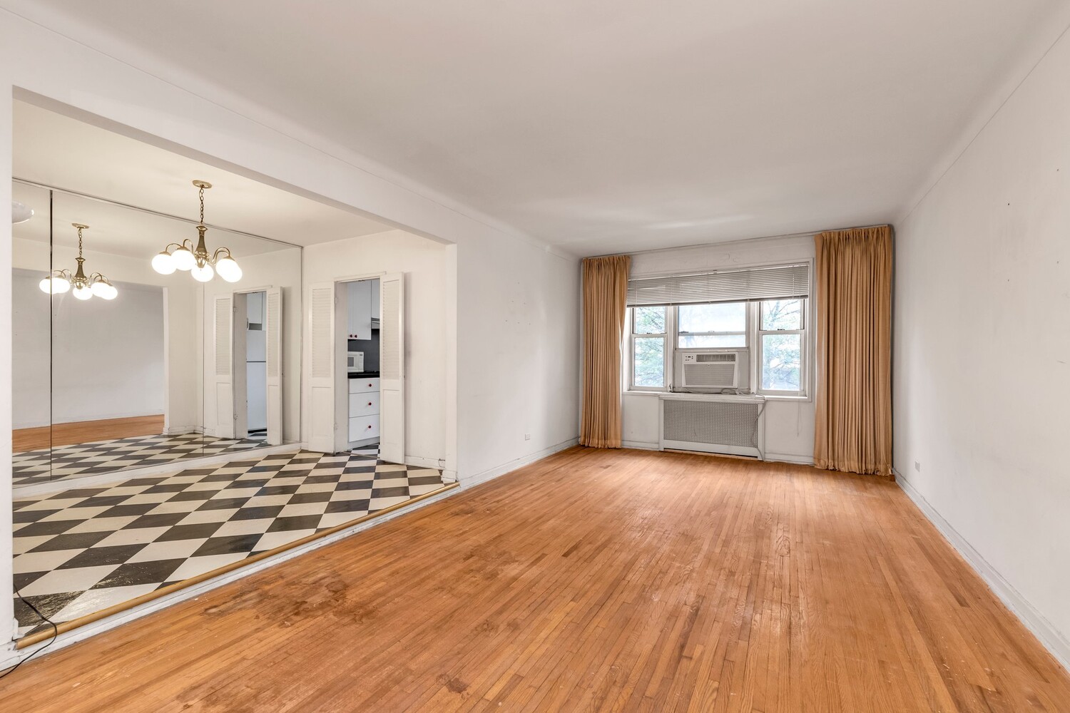 Vacant living room with hardwood floors, mirrored wall, checkerboard entry floor, and window air conditioner.