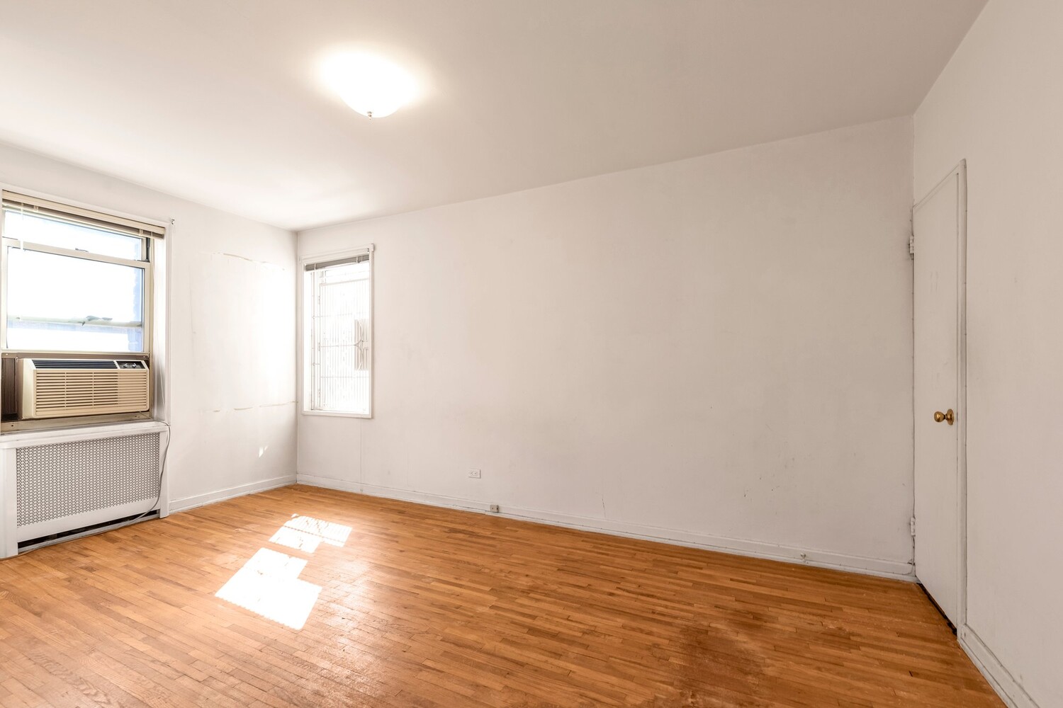 Empty apartment room with hardwood floors, two windows, and sunlight across the floor.