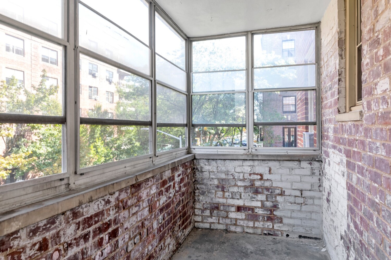 Enclosed sunroom with wraparound windows, exposed brick walls, and a bare concrete floor.