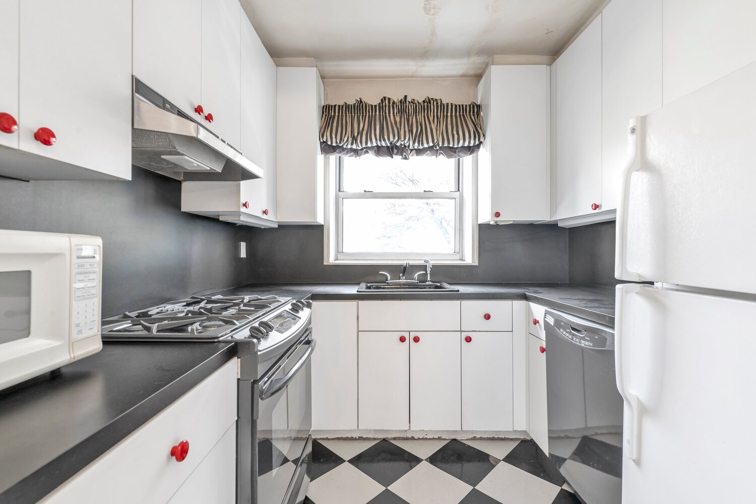 Compact kitchen with white cabinets, red knobs, dark counters, stainless steel appliances, and checkerboard flooring.