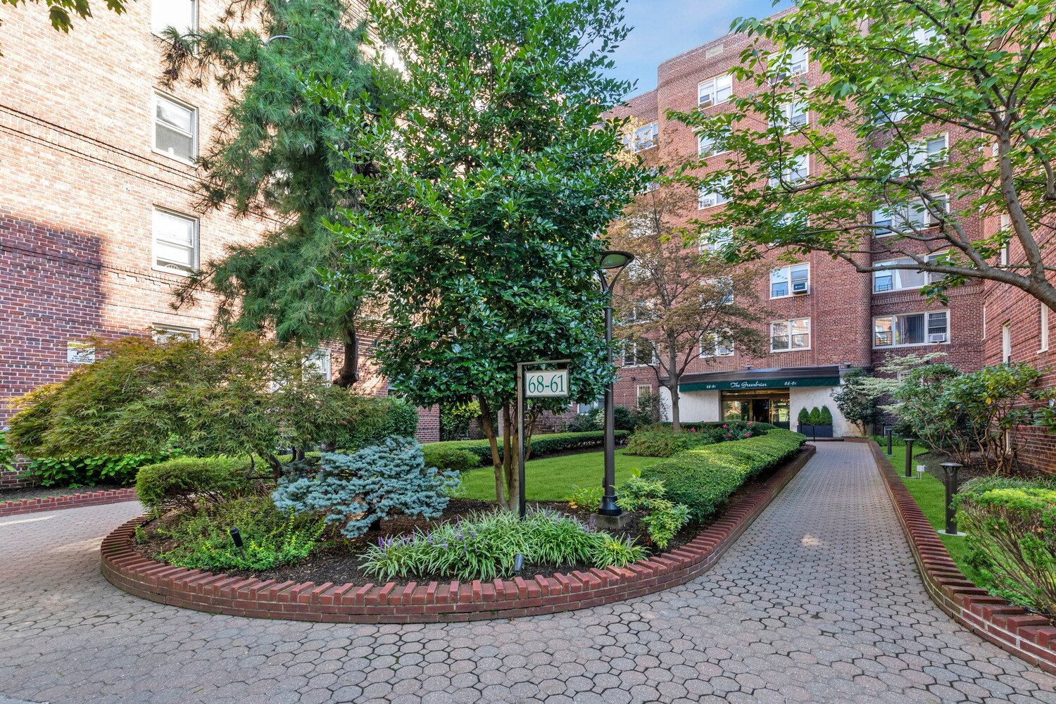 Apartment building entrance framed by landscaped gardens, mature trees, and a paved courtyard.