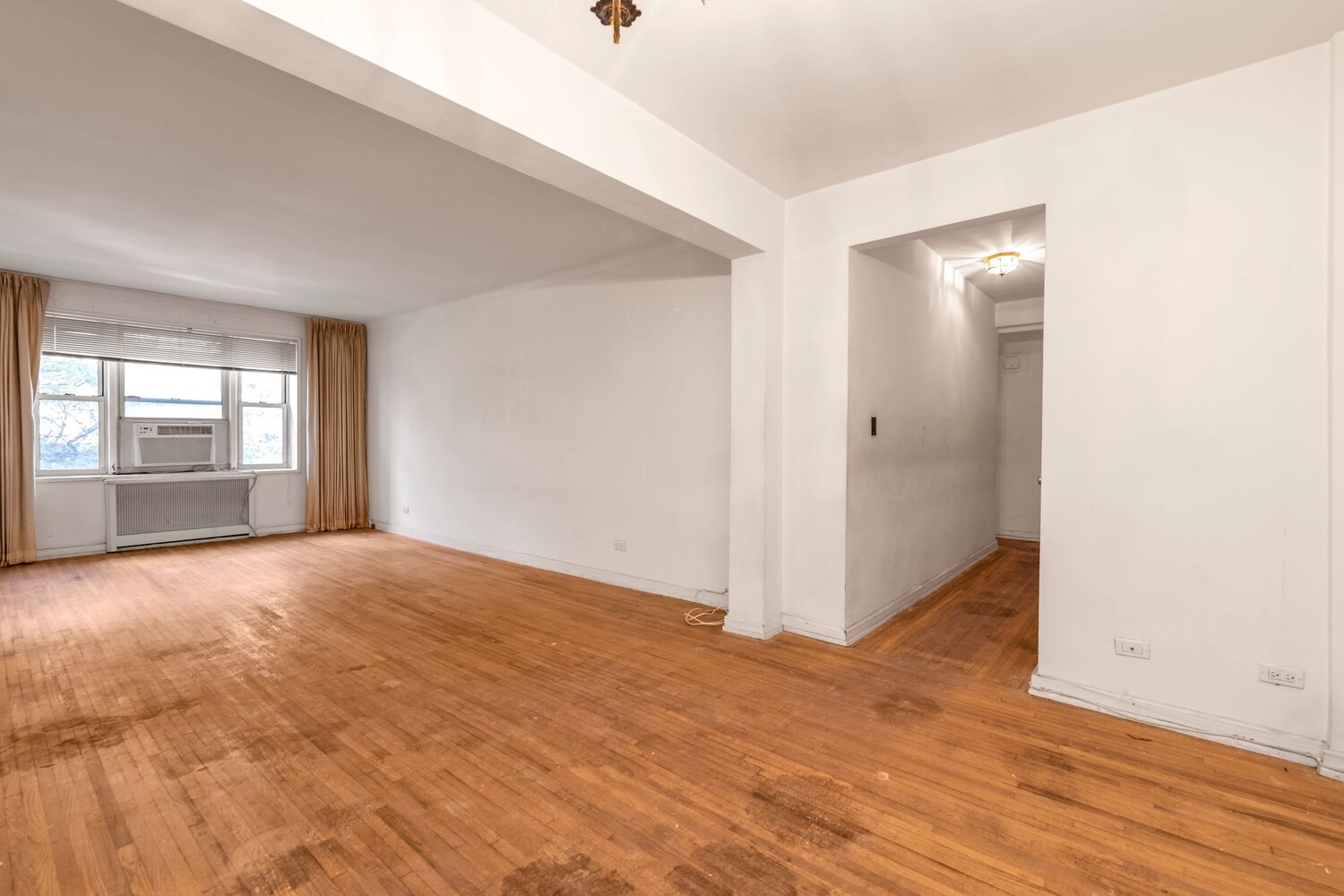 Empty living room with hardwood floors, a curtained window, and a window air conditioner.