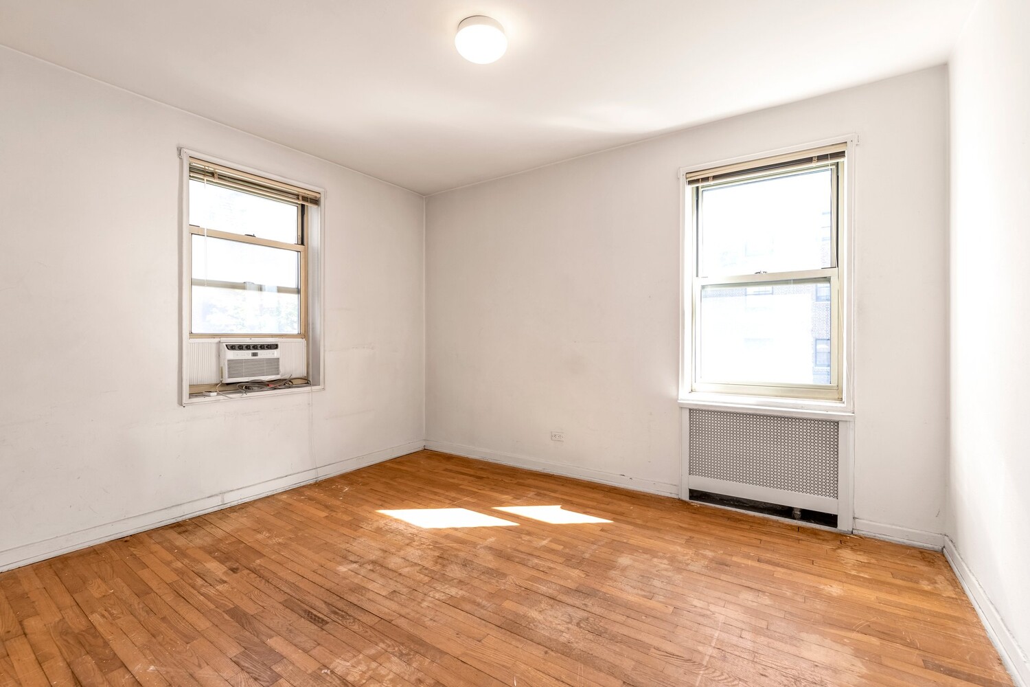 Vacant apartment room with hardwood floors, two windows, and a window air conditioner.