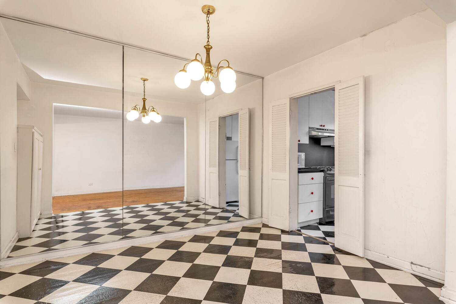 Empty dining area with checkerboard tile, mirrored wall, brass chandelier, and kitchen doorway.