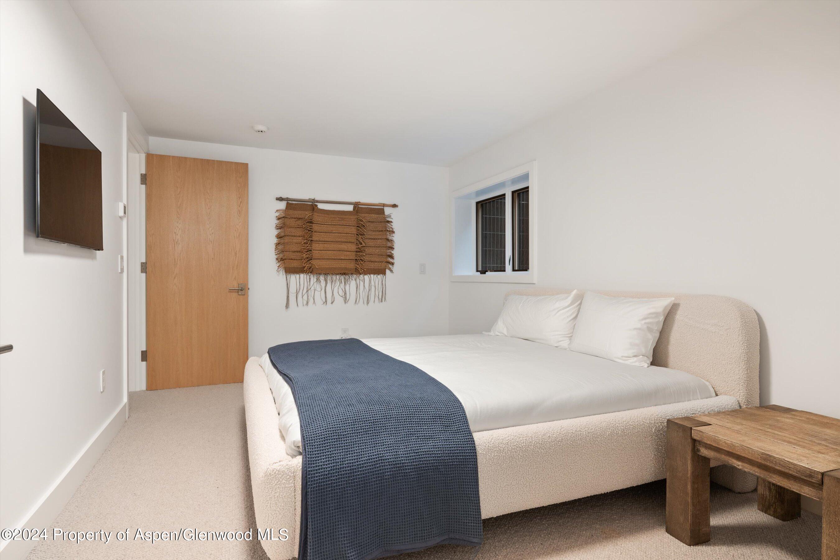 Bedroom with a cream upholstered bed, blue coverlet, wall-mounted TV, and woven wall hanging.