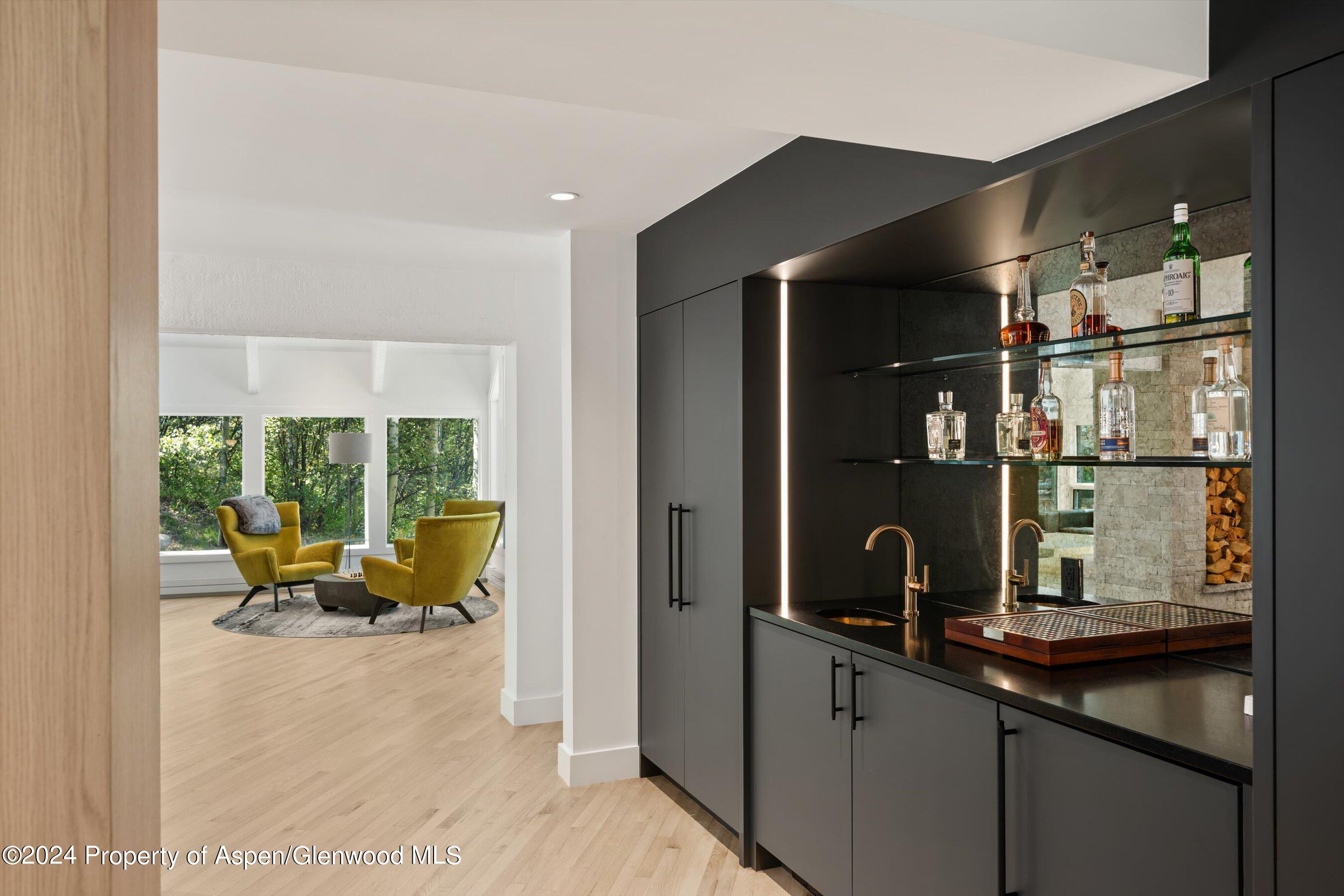 Built-in wet bar with glass liquor shelves beside a bright sitting area with mustard-yellow chairs.