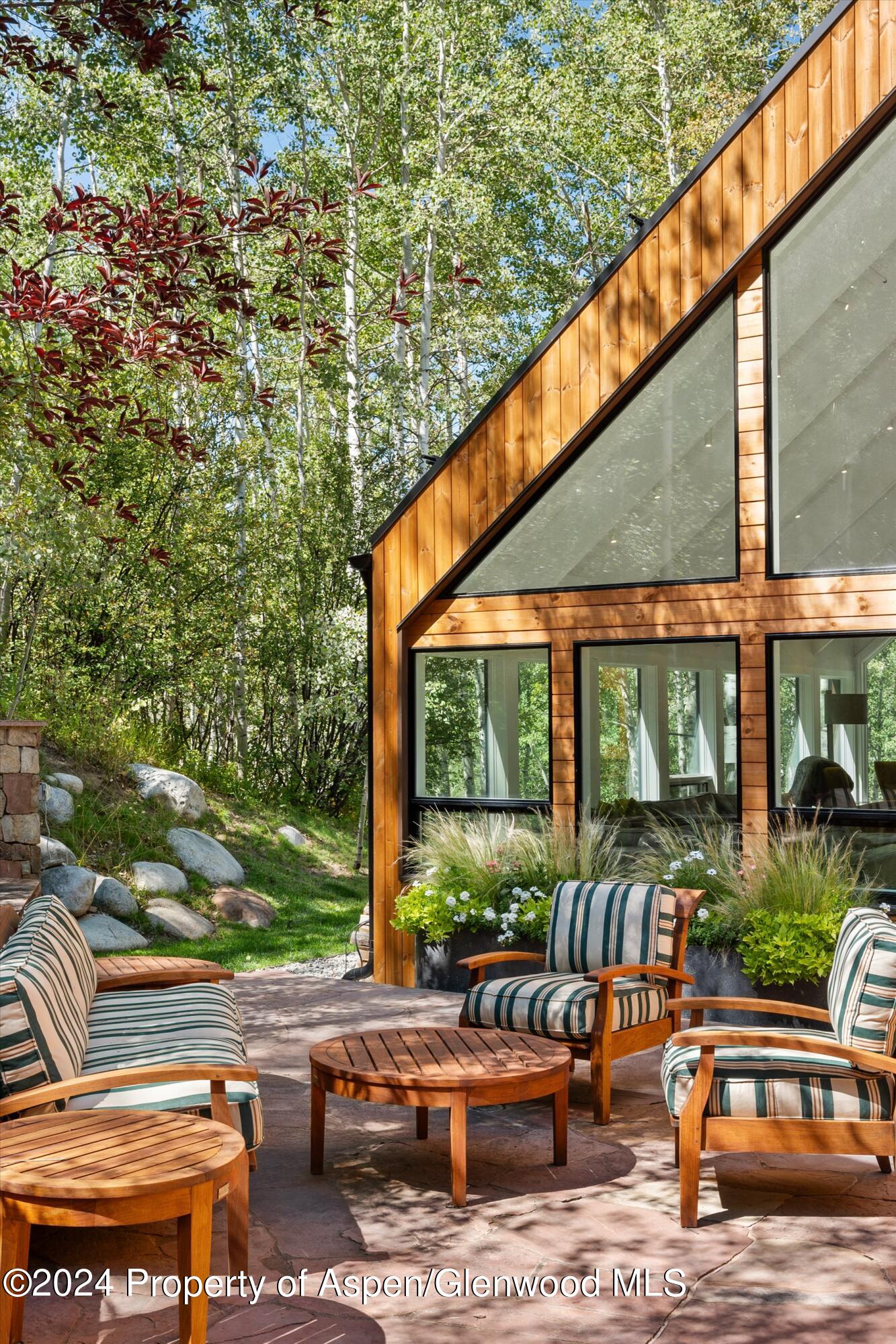 Outdoor patio with striped seating beside a wood-clad home, surrounded by trees.