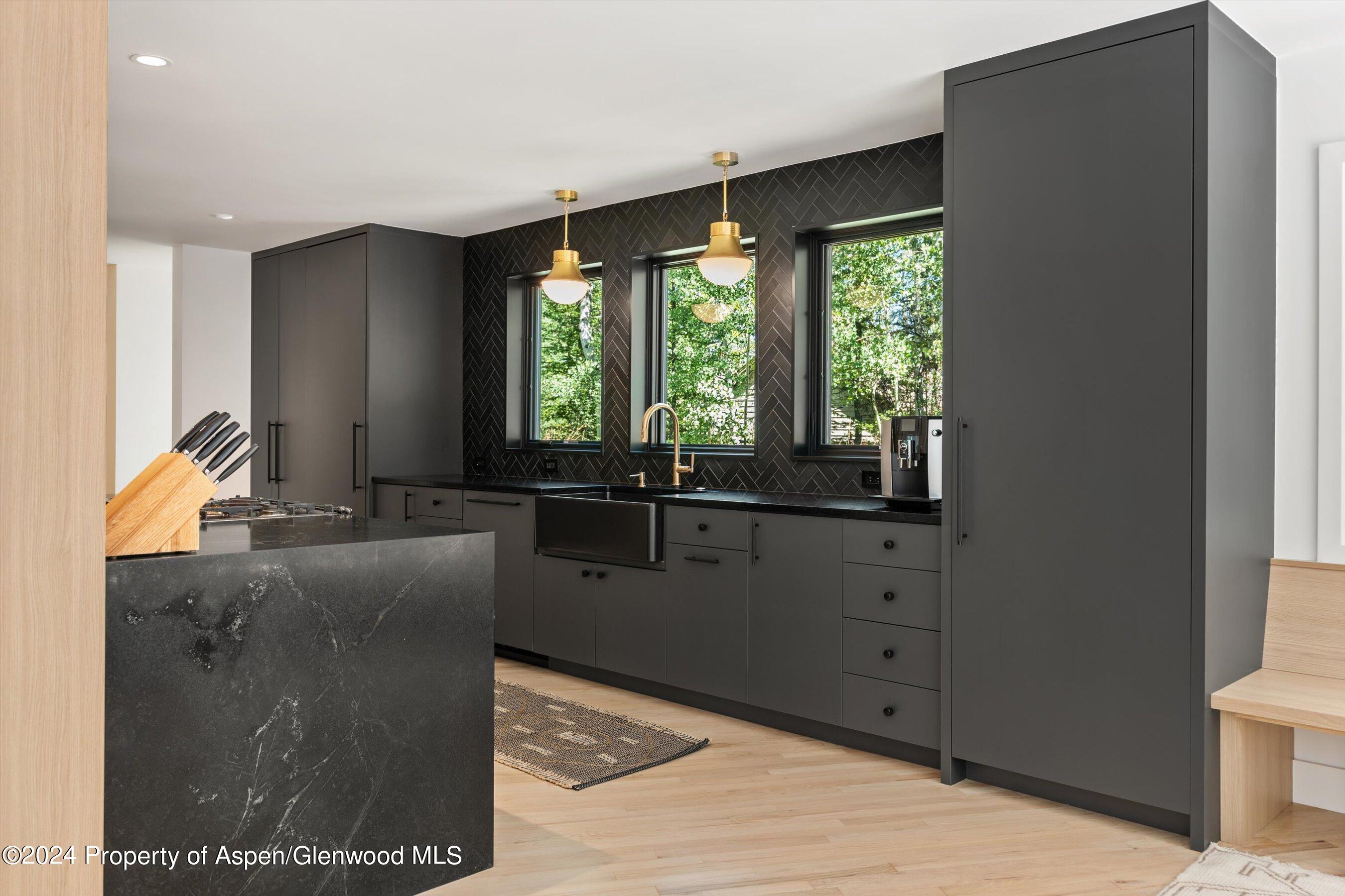 Modern kitchen with charcoal cabinetry, black stone island, herringbone backsplash, and three garden-facing windows.