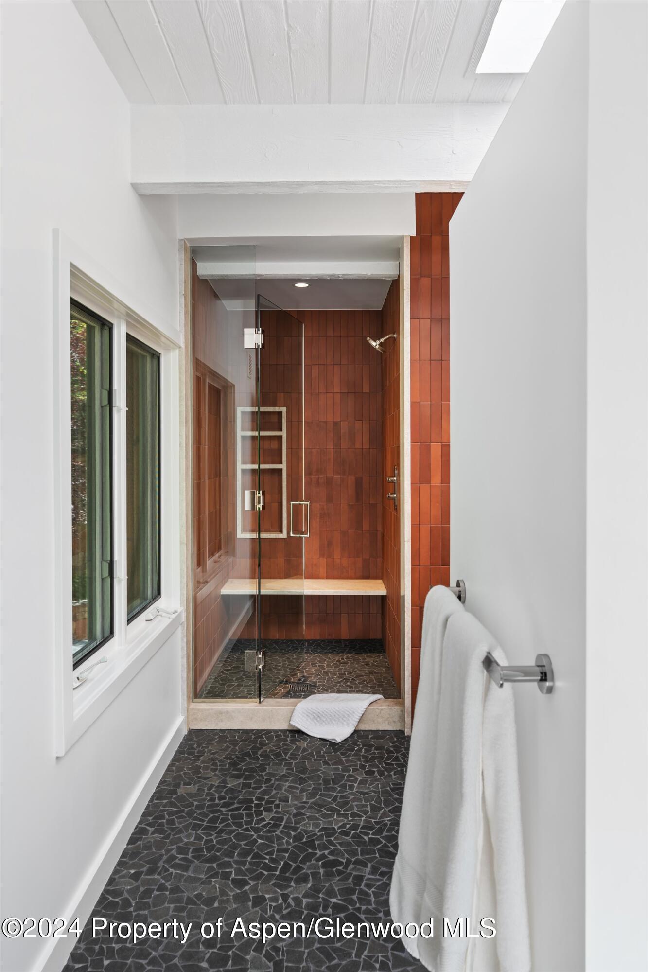 Walk-in shower with rust-colored tile, a glass door, and a built-in bench beside a window.