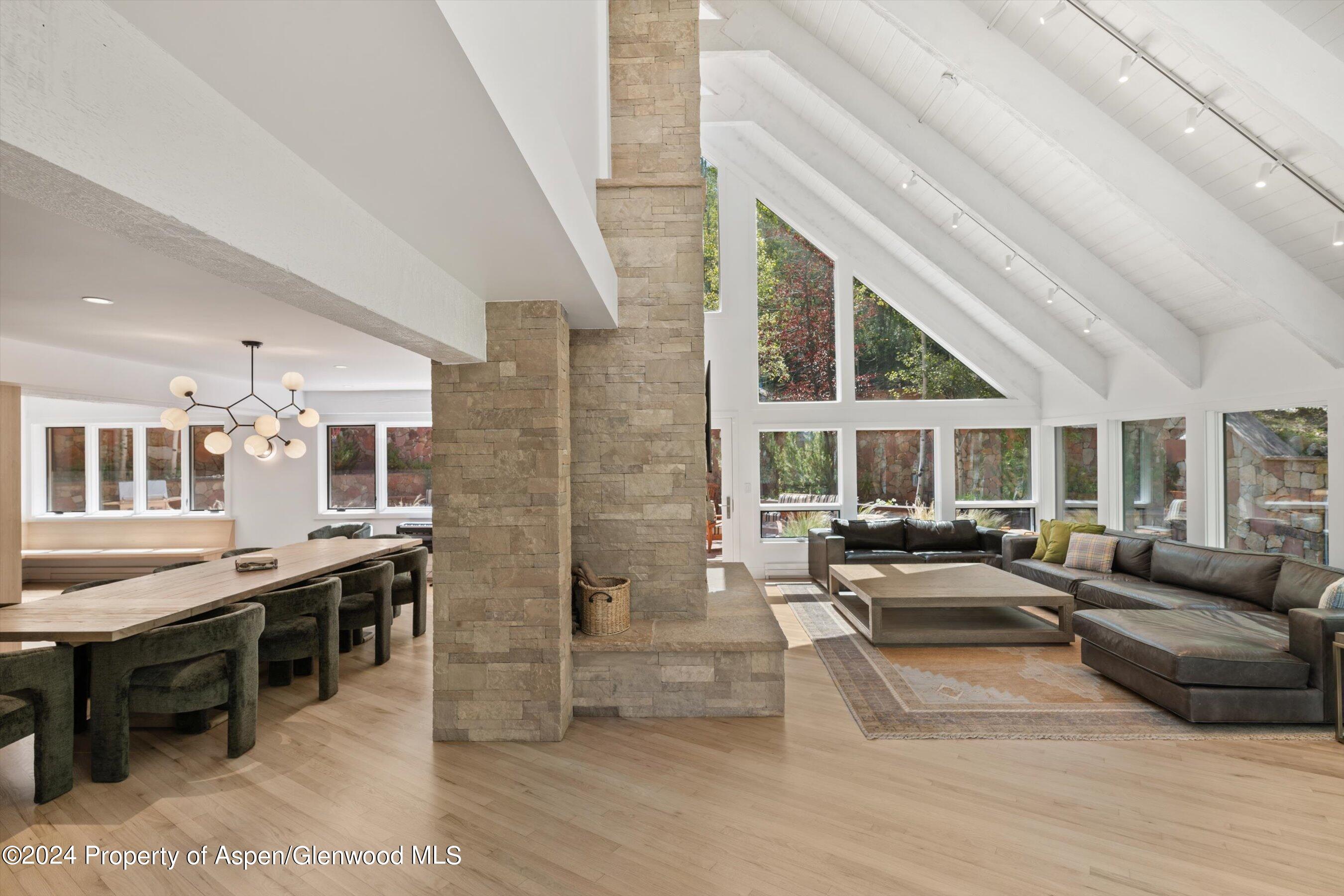 Open living and dining room with vaulted ceilings, a stone fireplace, and expansive windows.