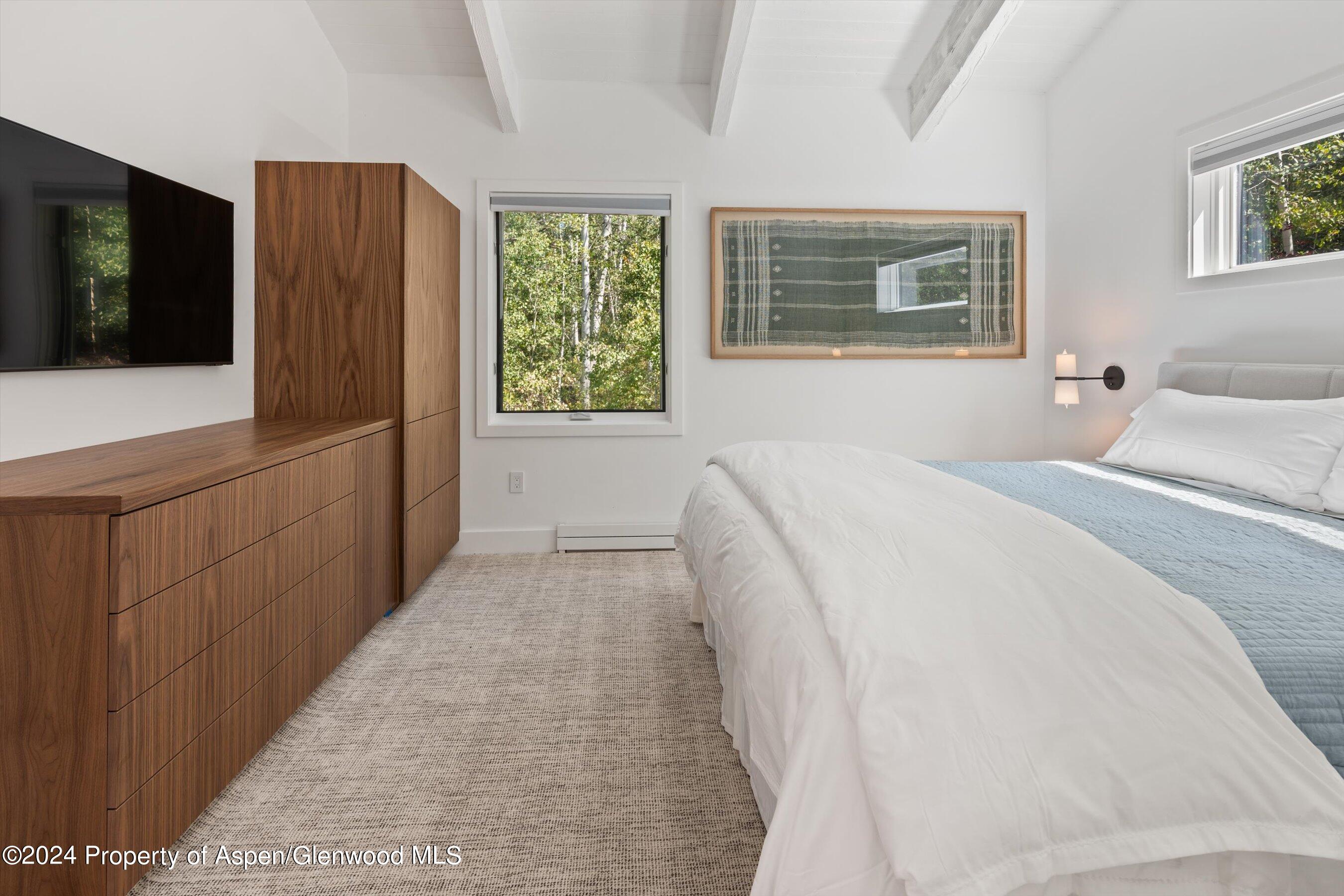 Bedroom with a white bed, walnut cabinetry, and windows overlooking leafy trees.