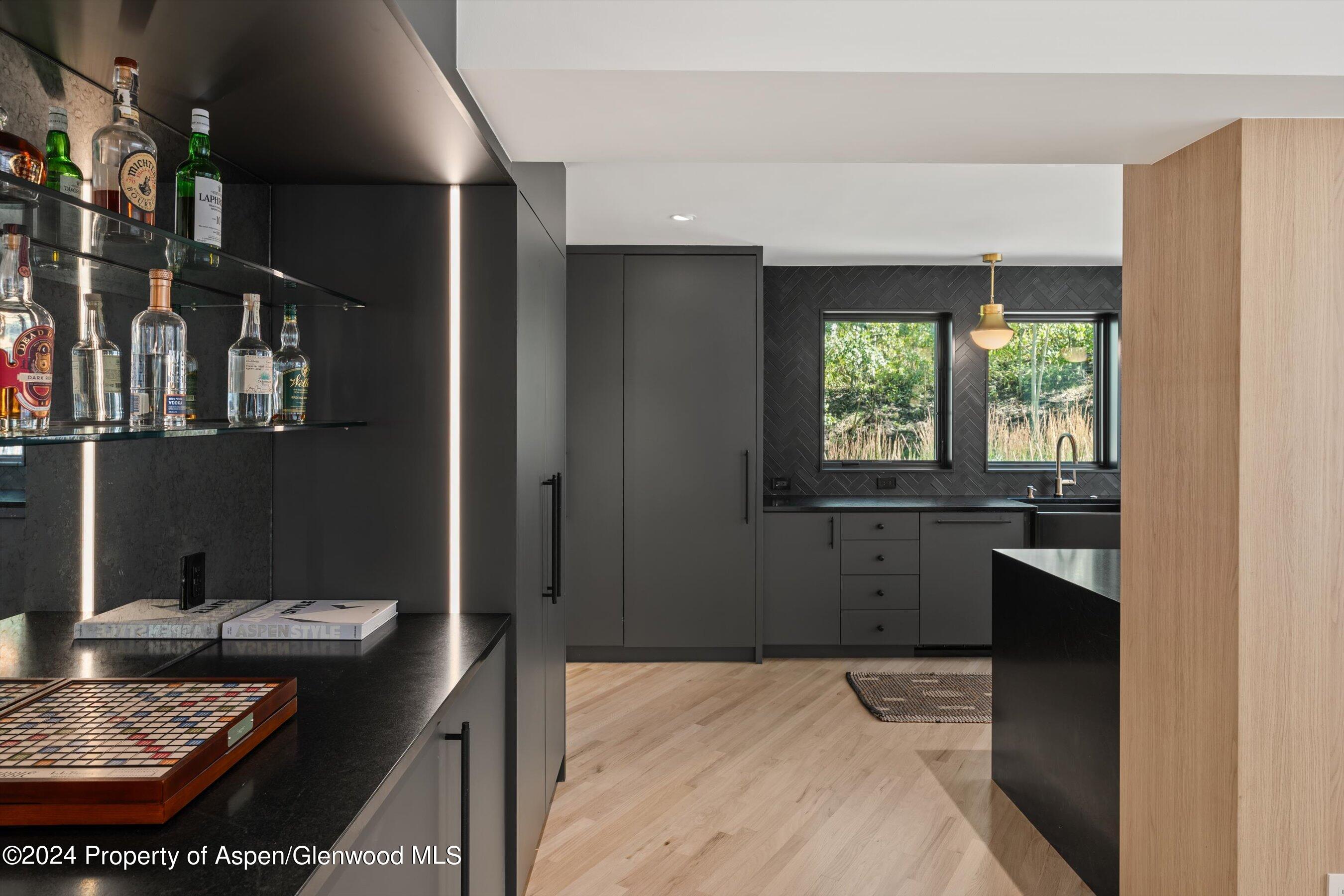 Modern kitchen with dark cabinetry, a bottle display, and windows overlooking greenery.
