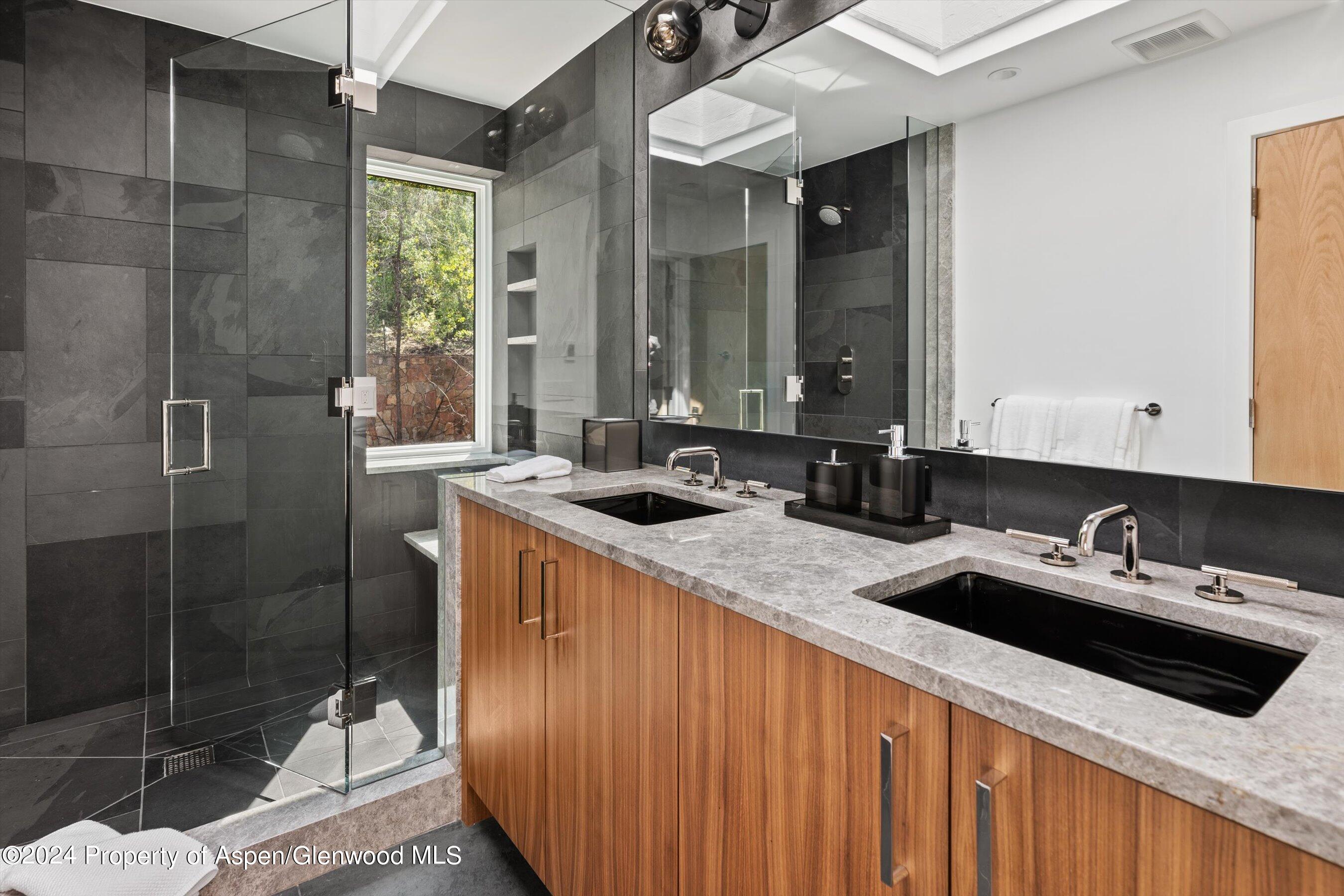 Bathroom with a glass shower, dark tile, and a double wood vanity beneath a large mirror.