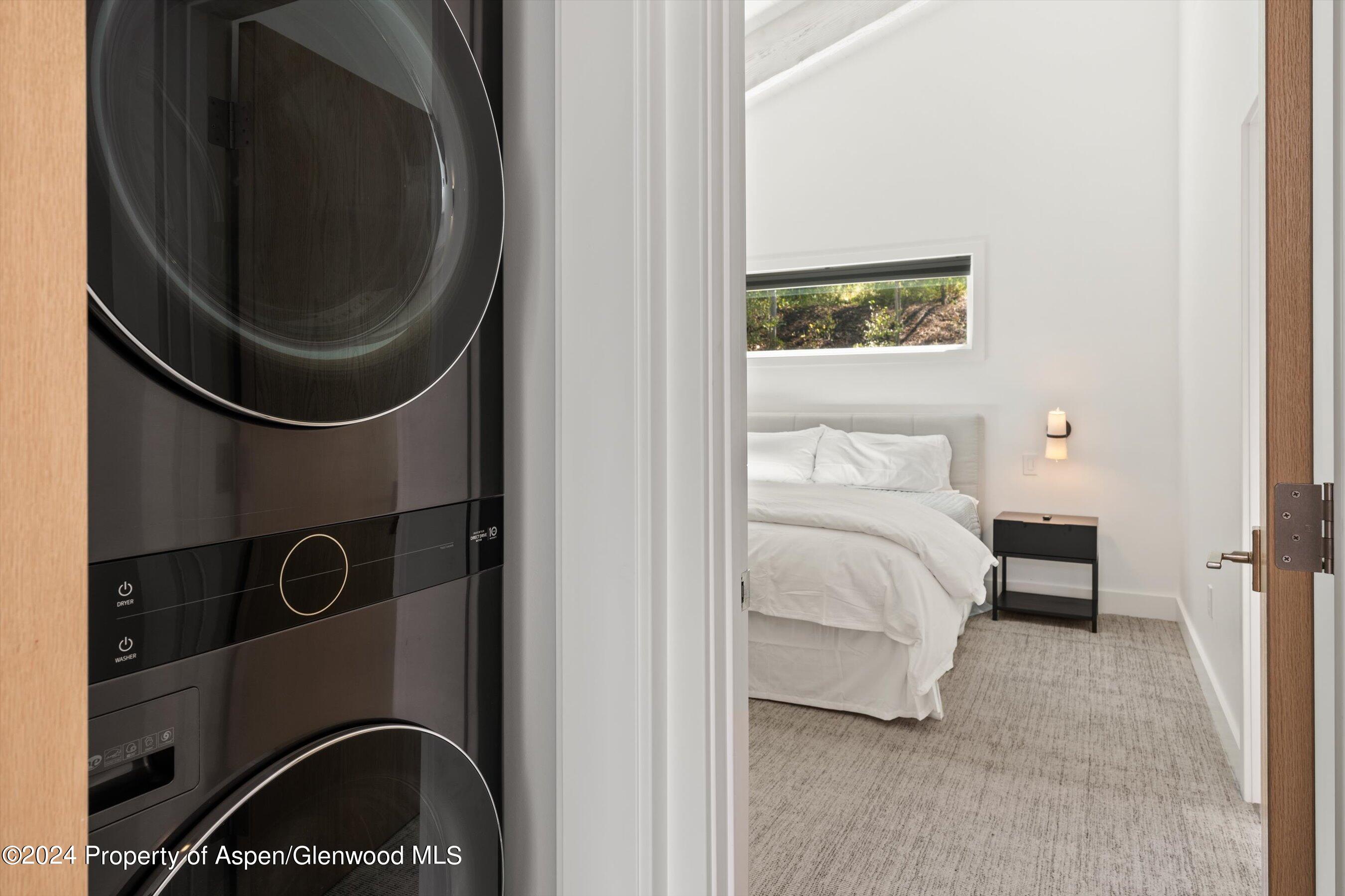 Bedroom with white bedding and a narrow window, viewed beside a stacked washer and dryer.