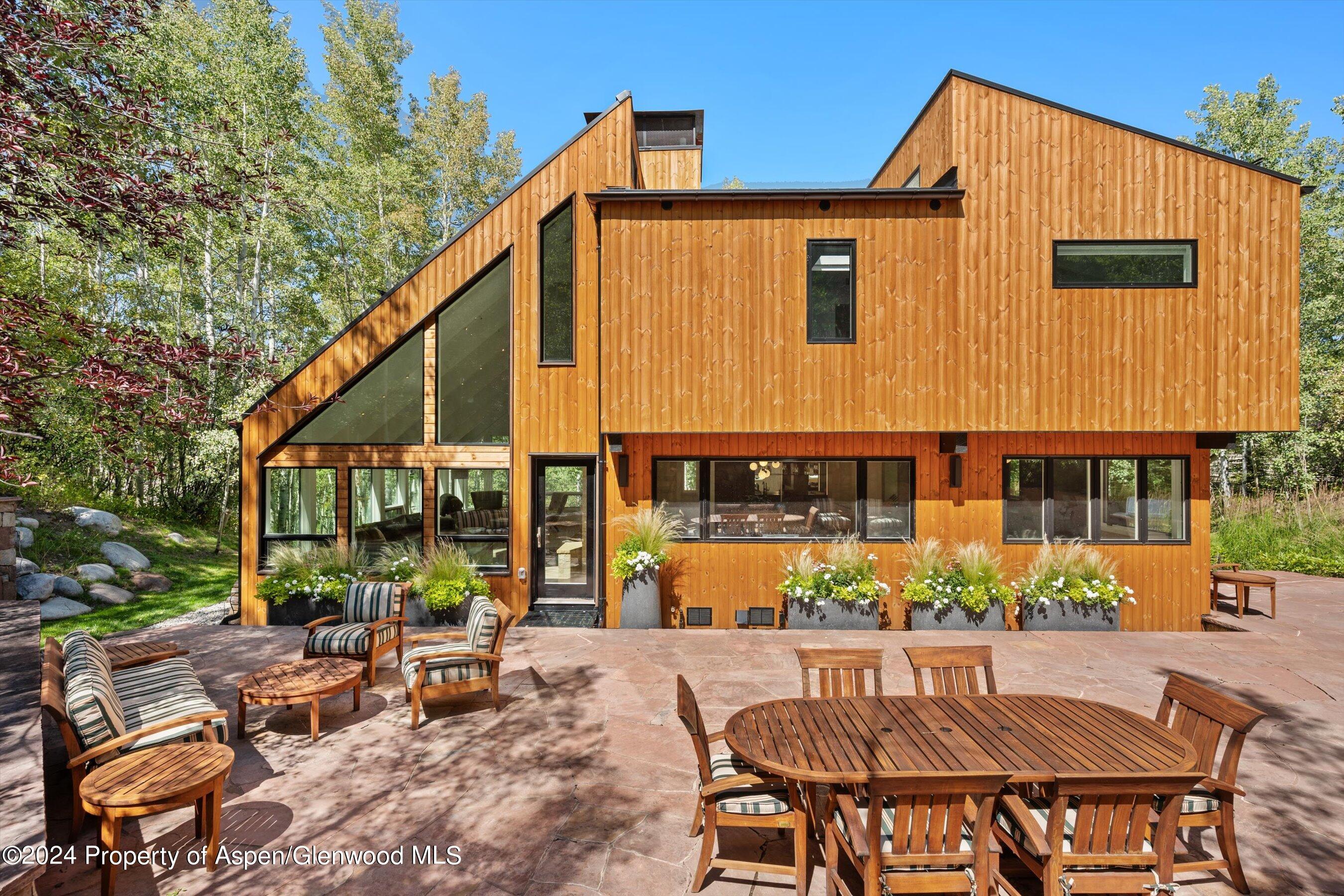 Modern wood-sided home with angular rooflines, expansive windows, and a furnished stone patio.
