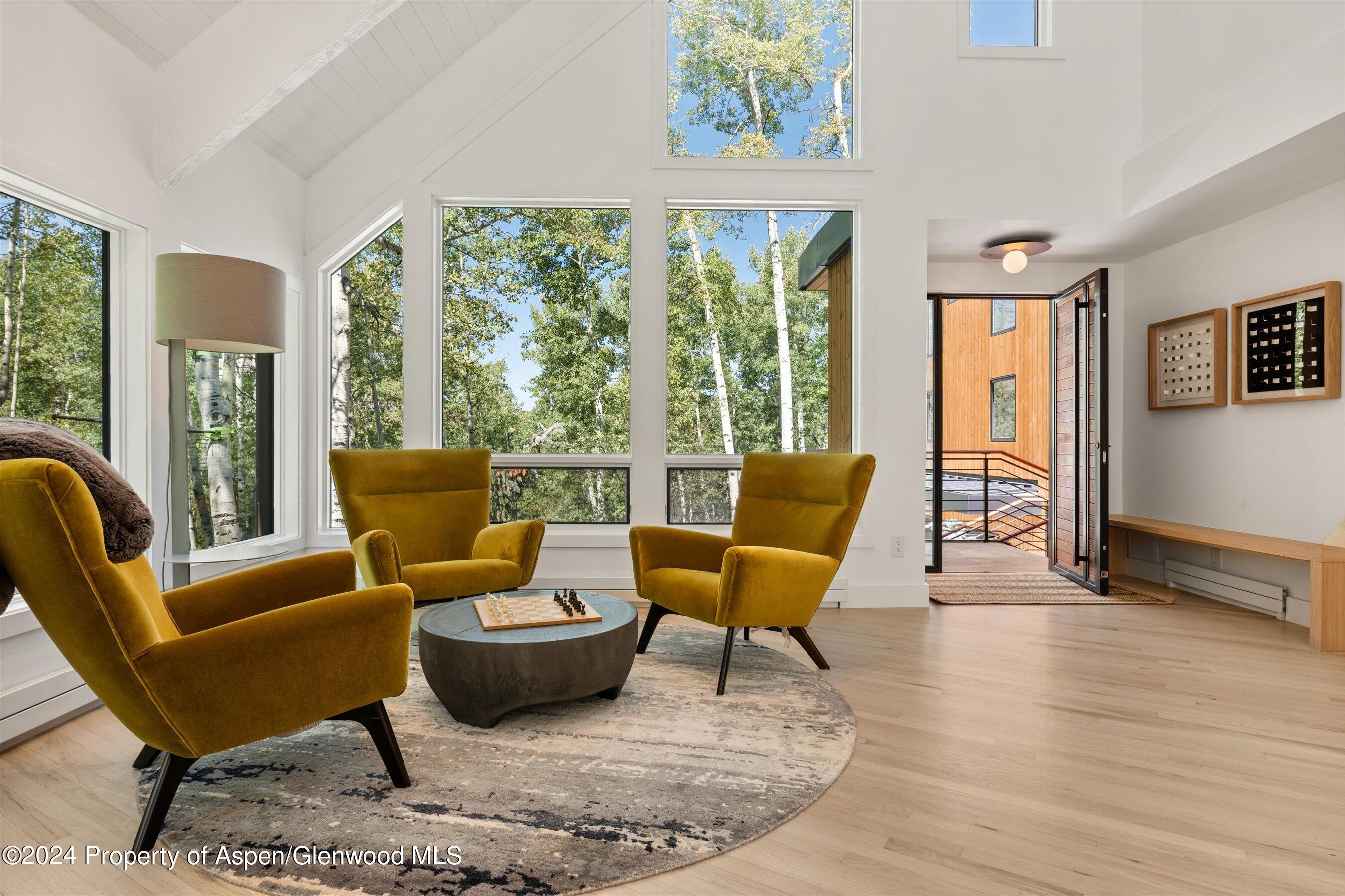 Living room with mustard armchairs, vaulted ceiling, and large windows overlooking leafy trees.