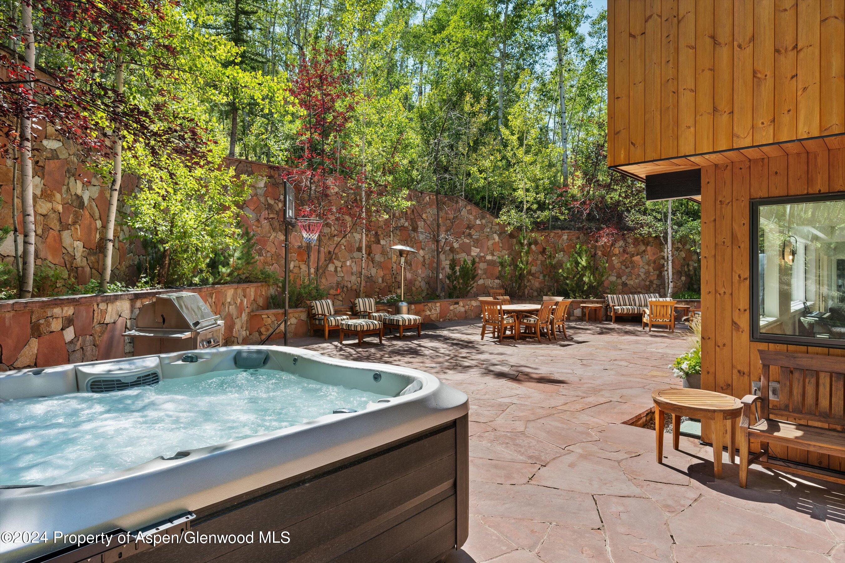 Stone patio with a bubbling hot tub, outdoor dining area, and basketball hoop beside a wood-clad home.