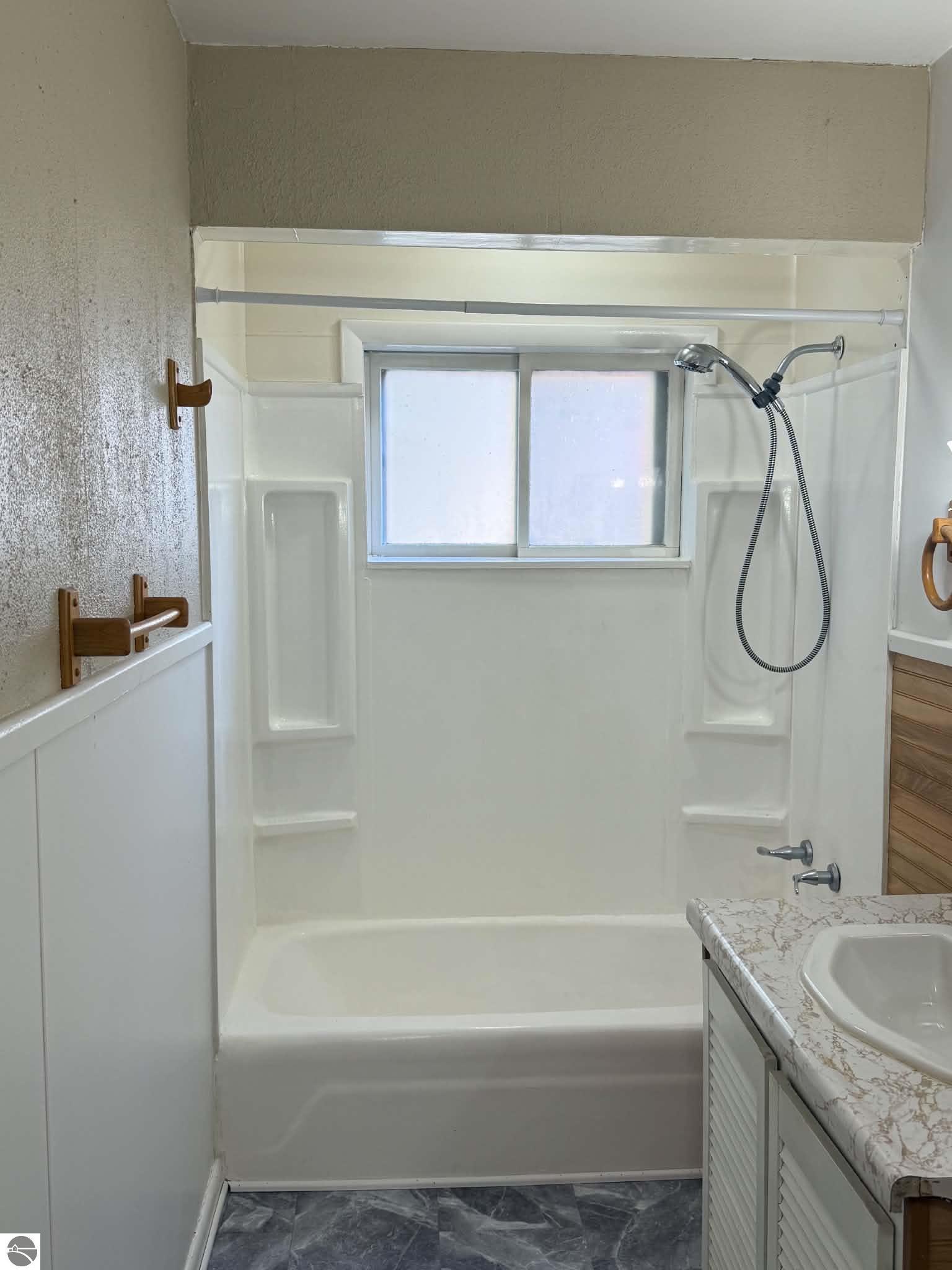 Bathroom with a white tub and shower beneath a frosted window, beside a marble-pattern vanity.