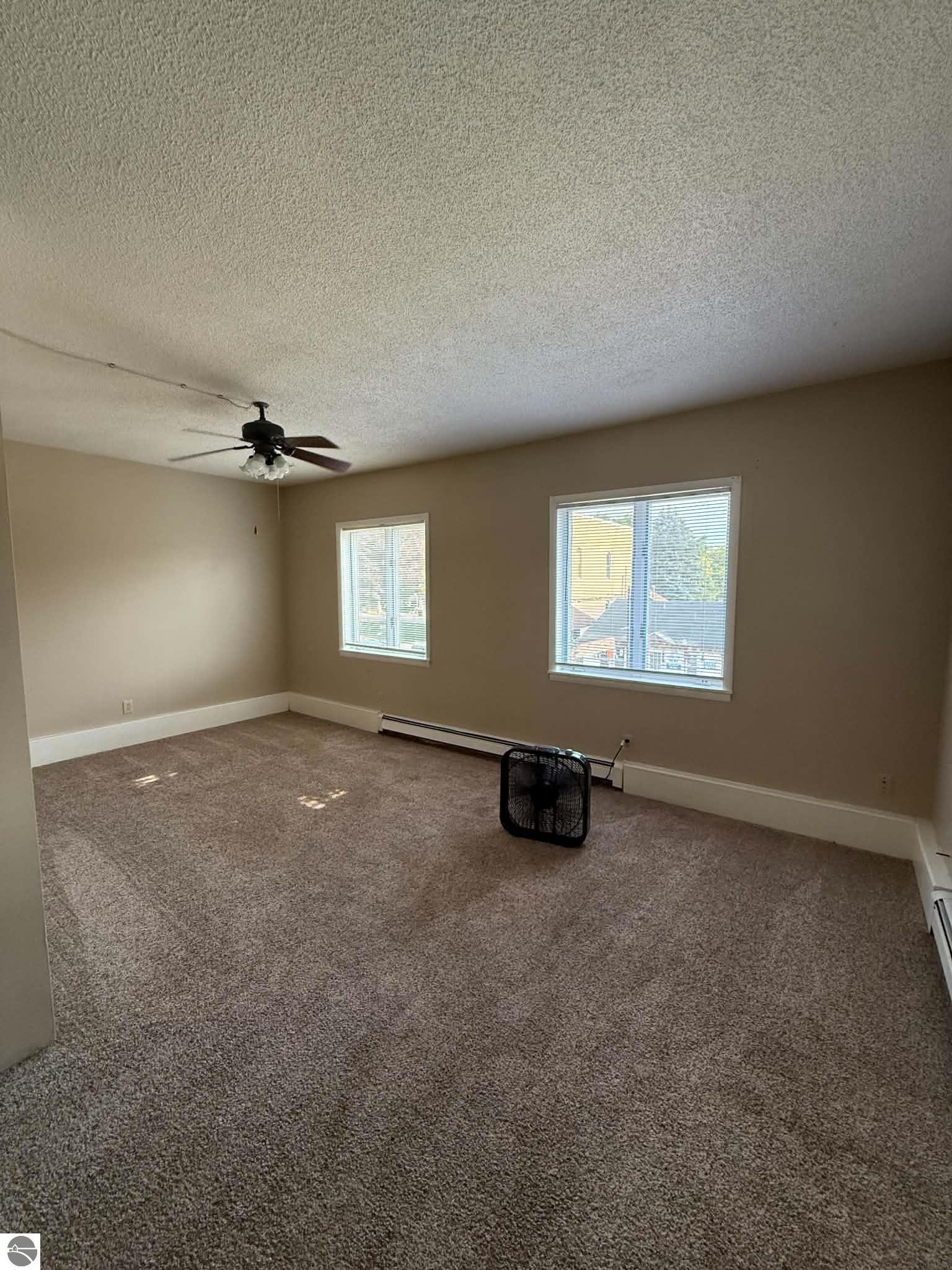 Empty carpeted room with two blinded windows, a ceiling fan, and baseboard heating.