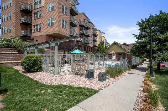Brick apartment building with balconies beside a fenced swimming pool and landscaped sidewalk.
