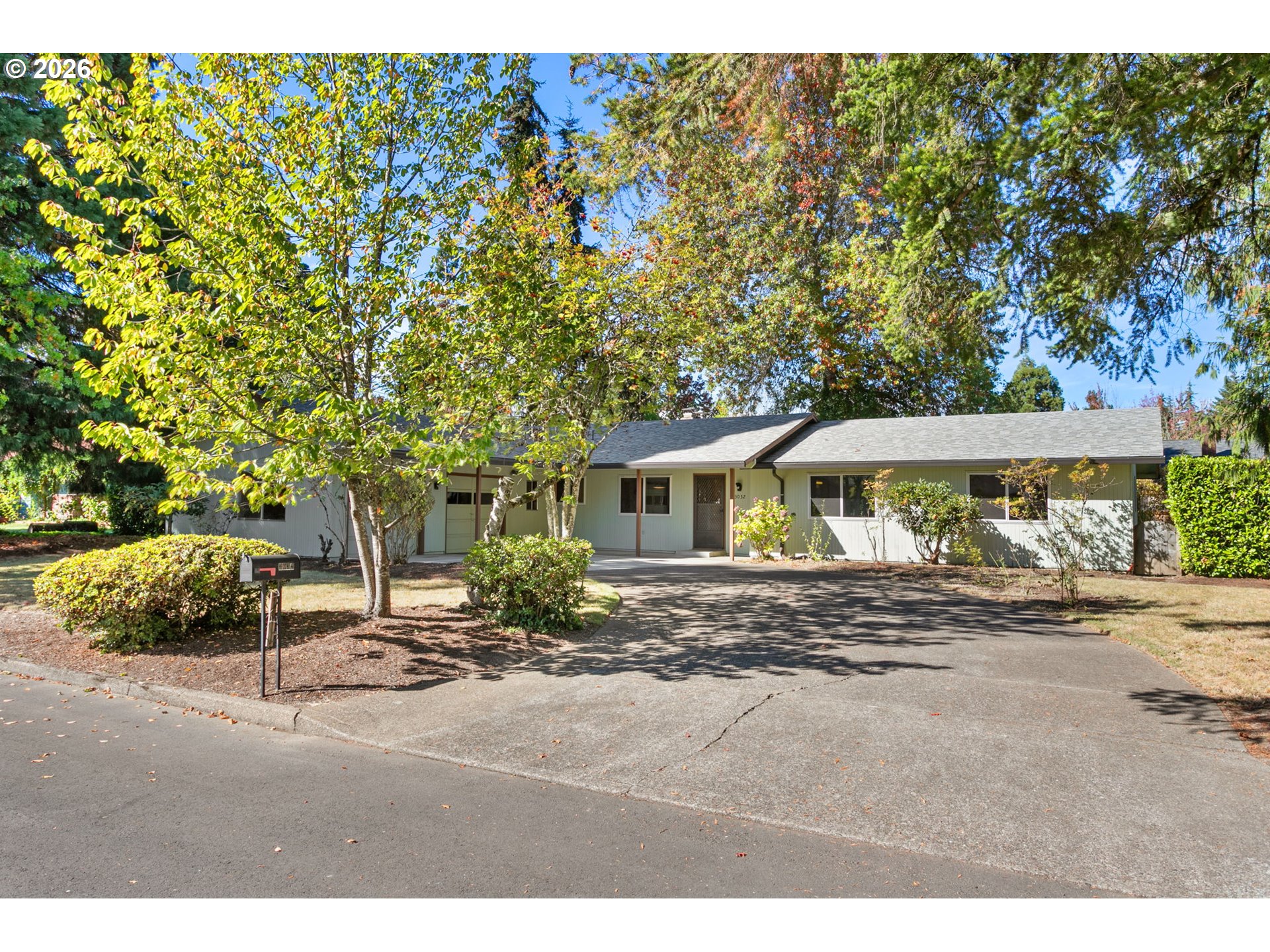 Single-story home with pale siding, a broad driveway, mature trees, and a landscaped front yard.