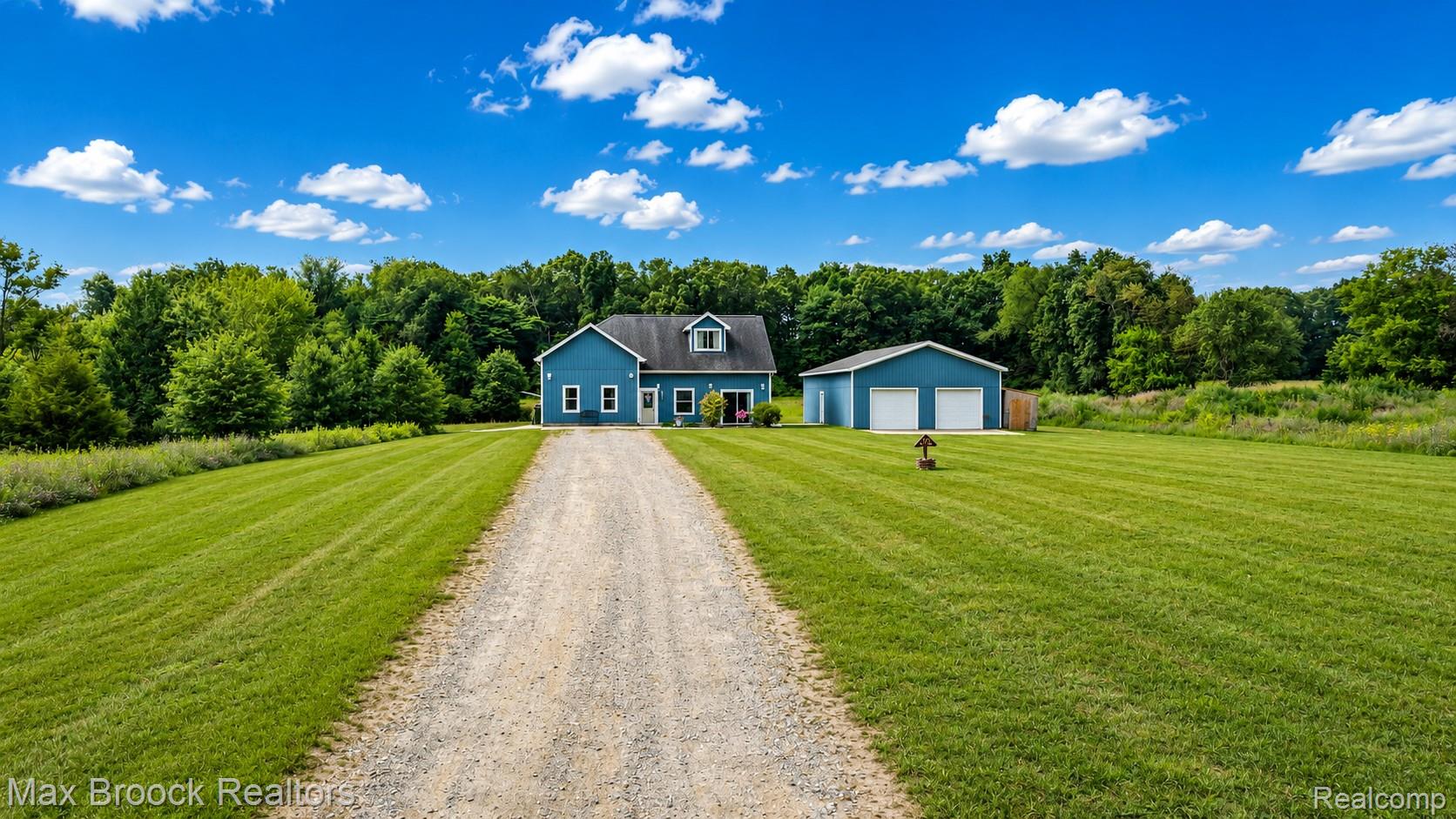 Blue two-story house and detached garage at the end of a gravel driveway, surrounded by a broad lawn and trees.