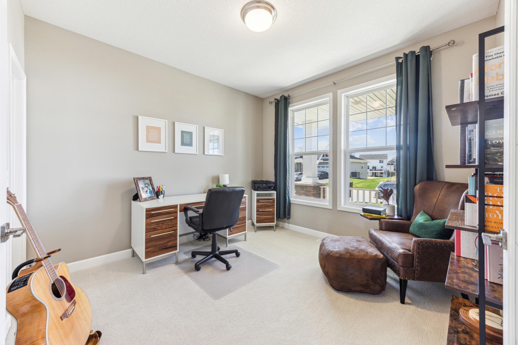 Home office with a desk and chair, large curtained windows, brown leather sofa, and acoustic guitar.