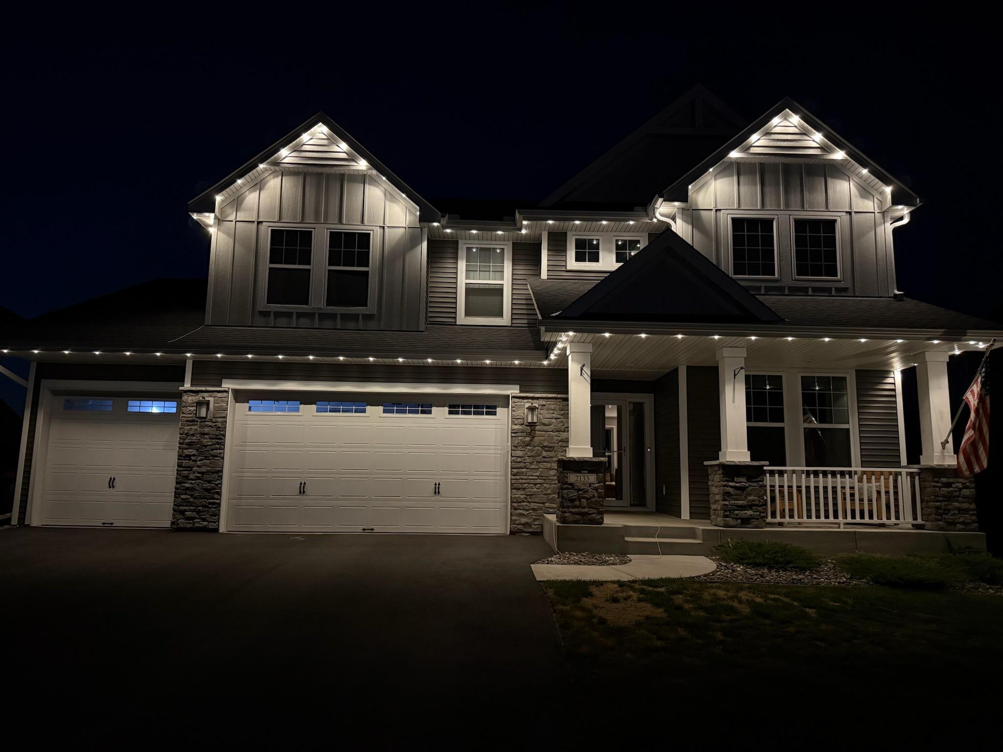 Two-story gray house at night with stone accents, a covered porch, and two garage doors.