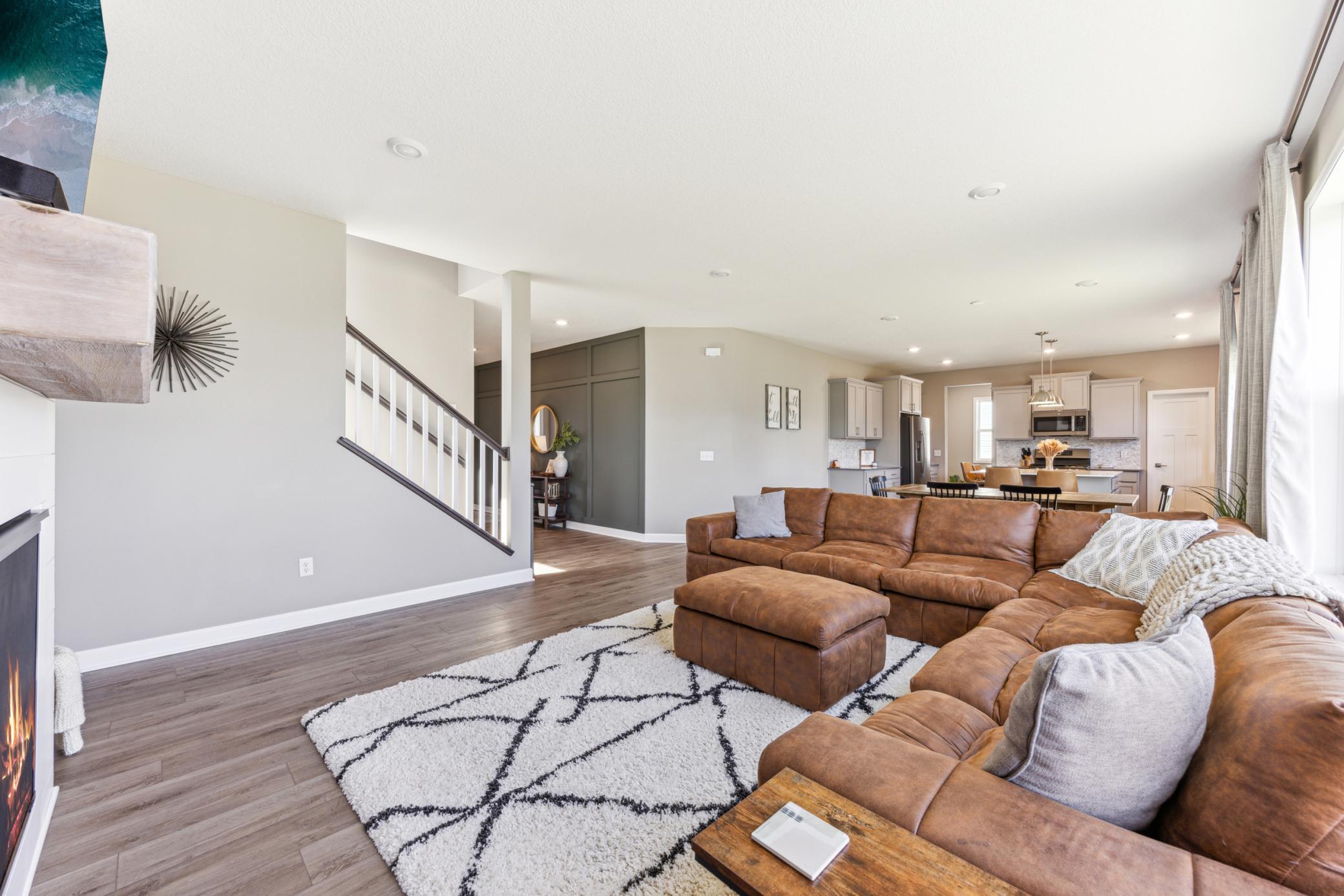 Open-plan living room with a brown leather sectional, geometric rug, staircase, and kitchen beyond.