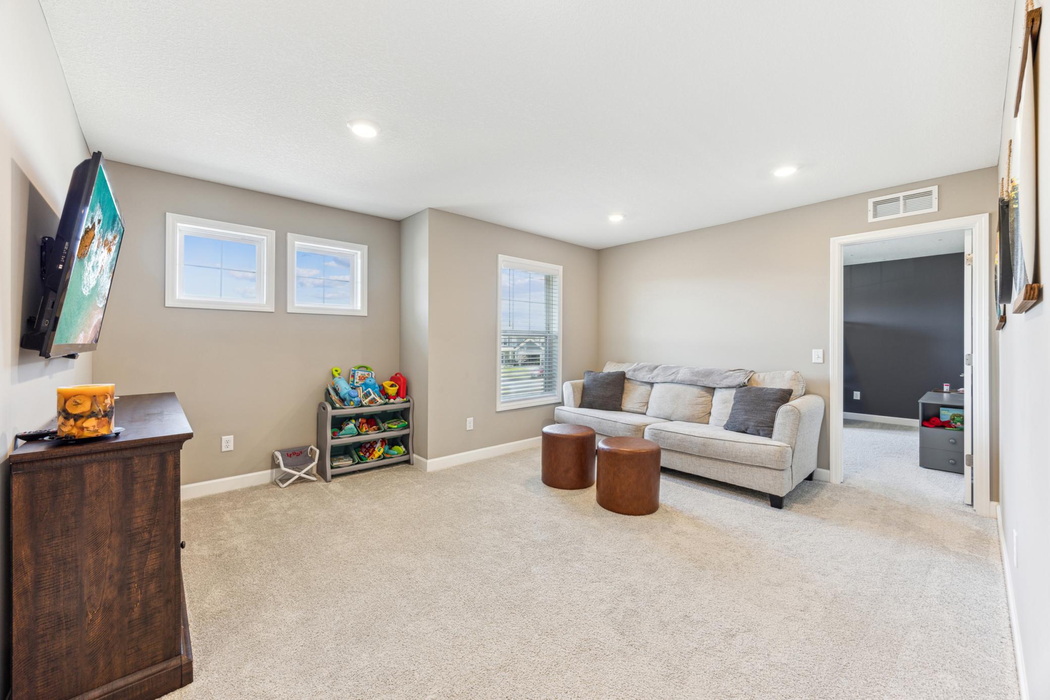Carpeted living room with a gray sofa, brown ottomans, wall-mounted TV, and children's toy shelf.