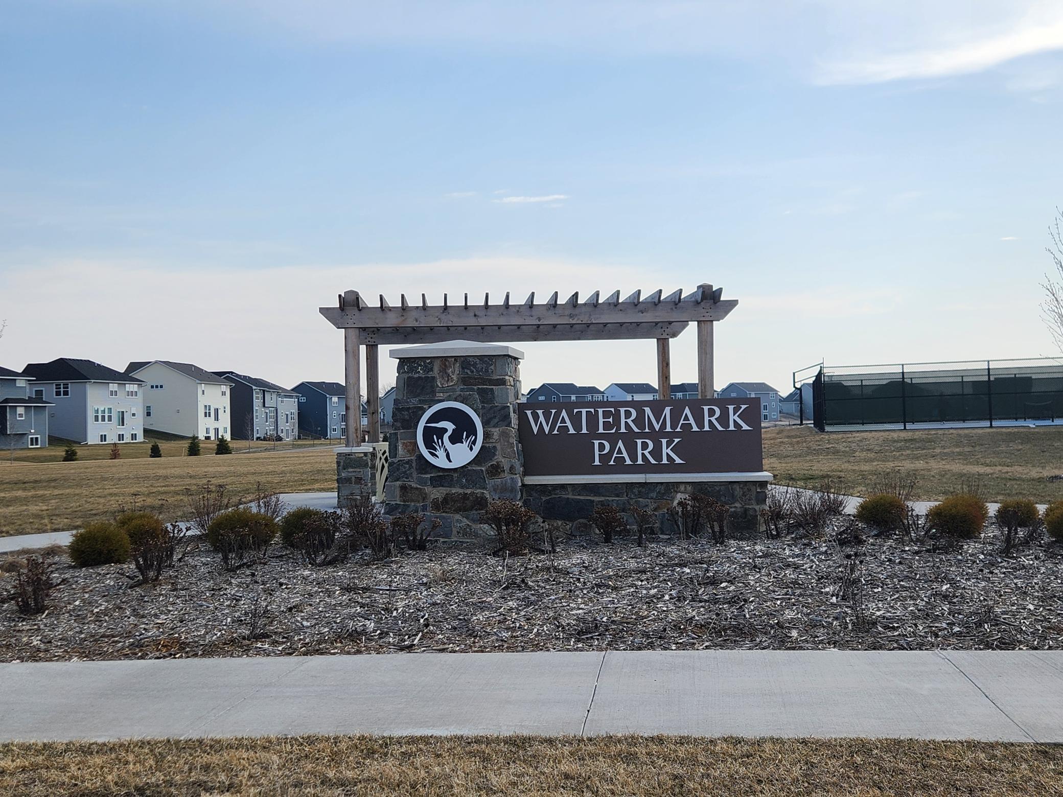 Watermark Park entrance sign beneath a wooden pergola, with homes and a fenced court behind it.