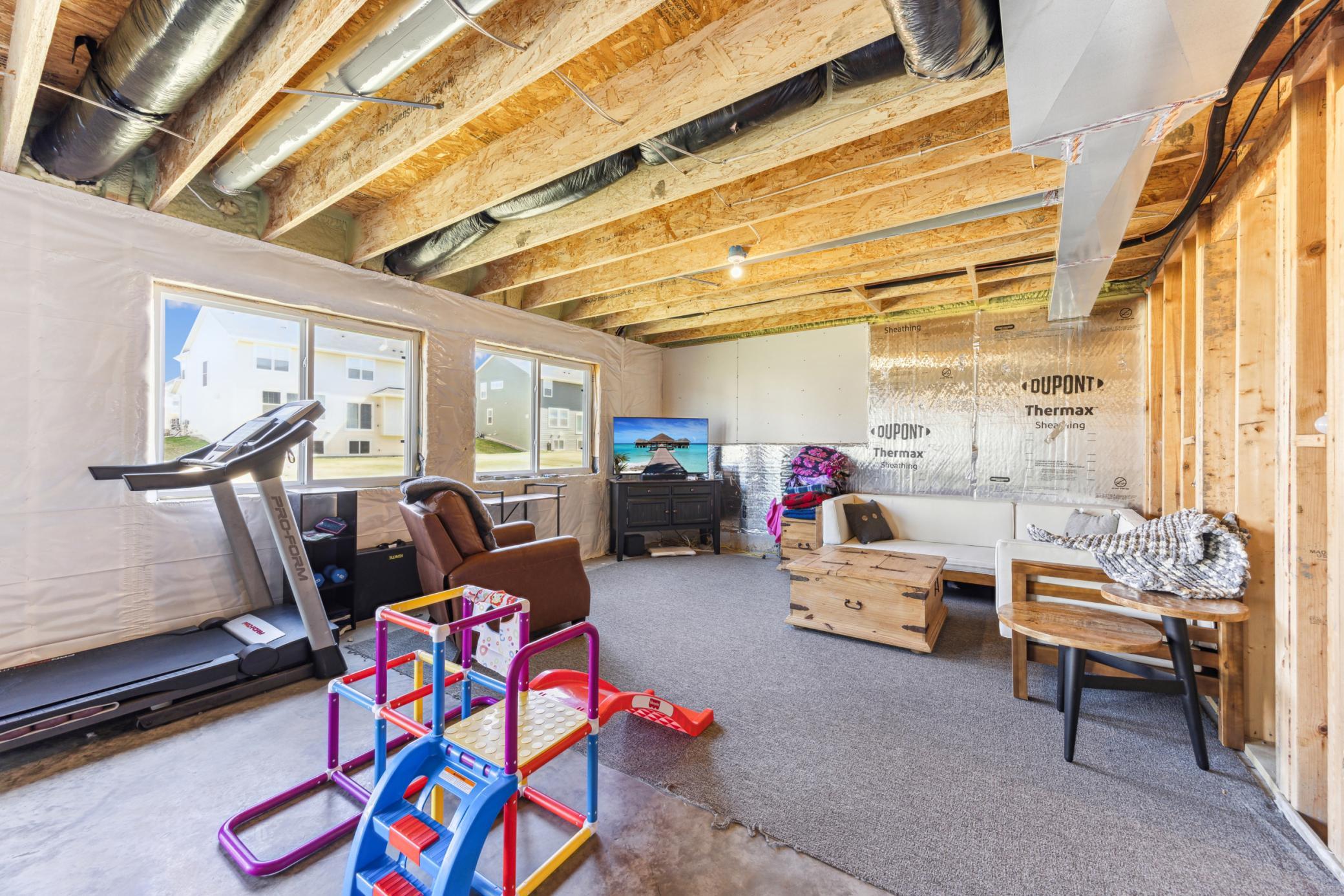 Partially unfinished basement recreation room with exposed ceiling framing, a treadmill, and a colorful children’s play structure.