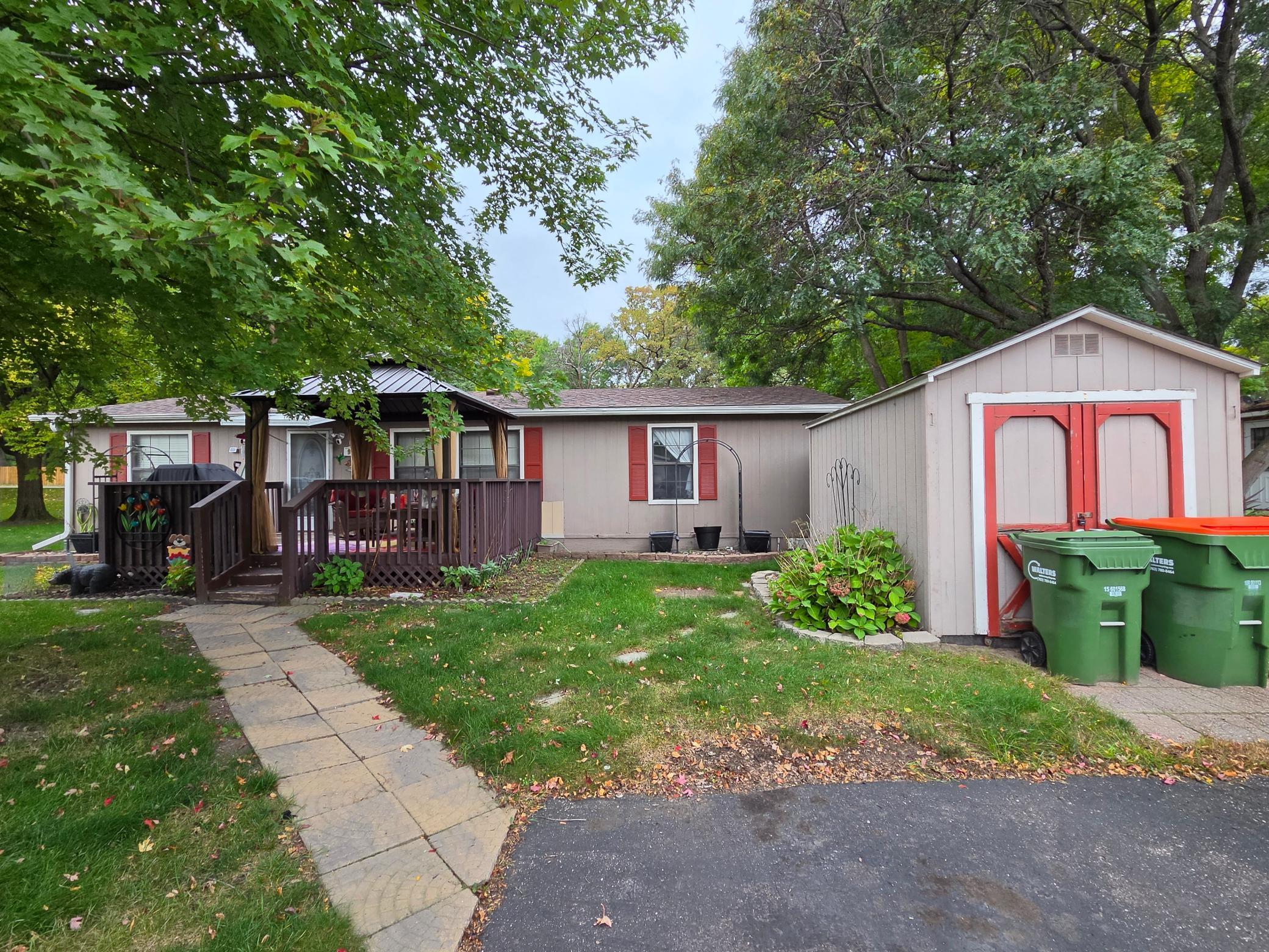 Single-story home with a covered porch, red shutters, and a detached shed beneath mature trees.