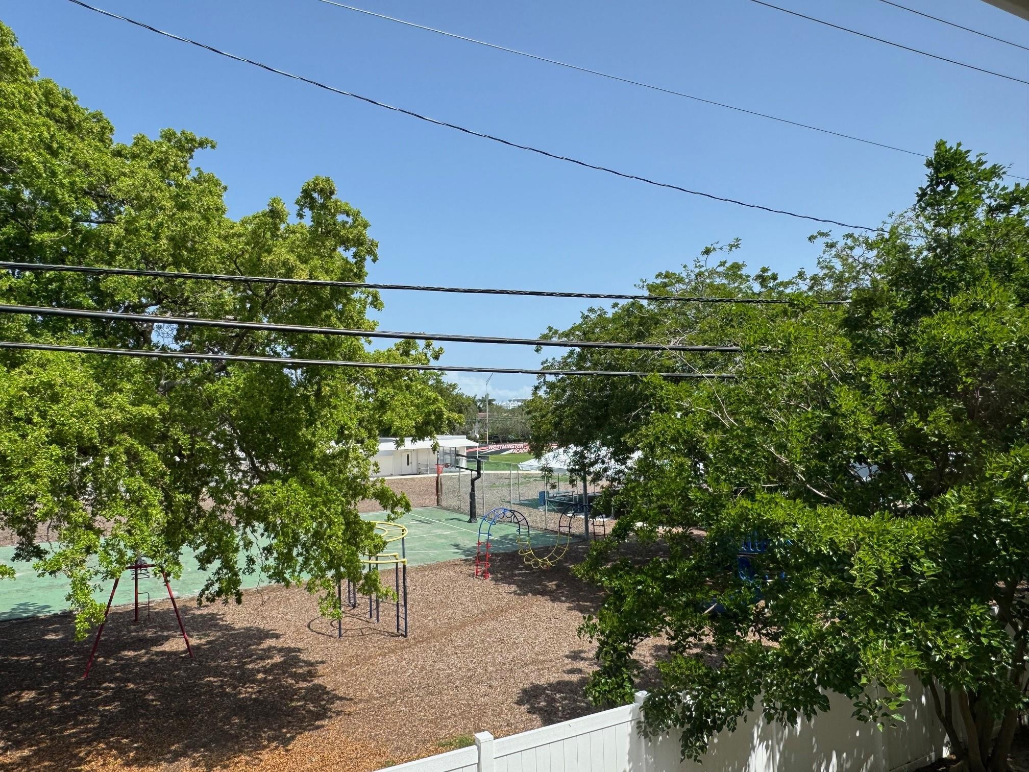 Tree-shaded playground with climbing equipment and a basketball court beyond a white fence.