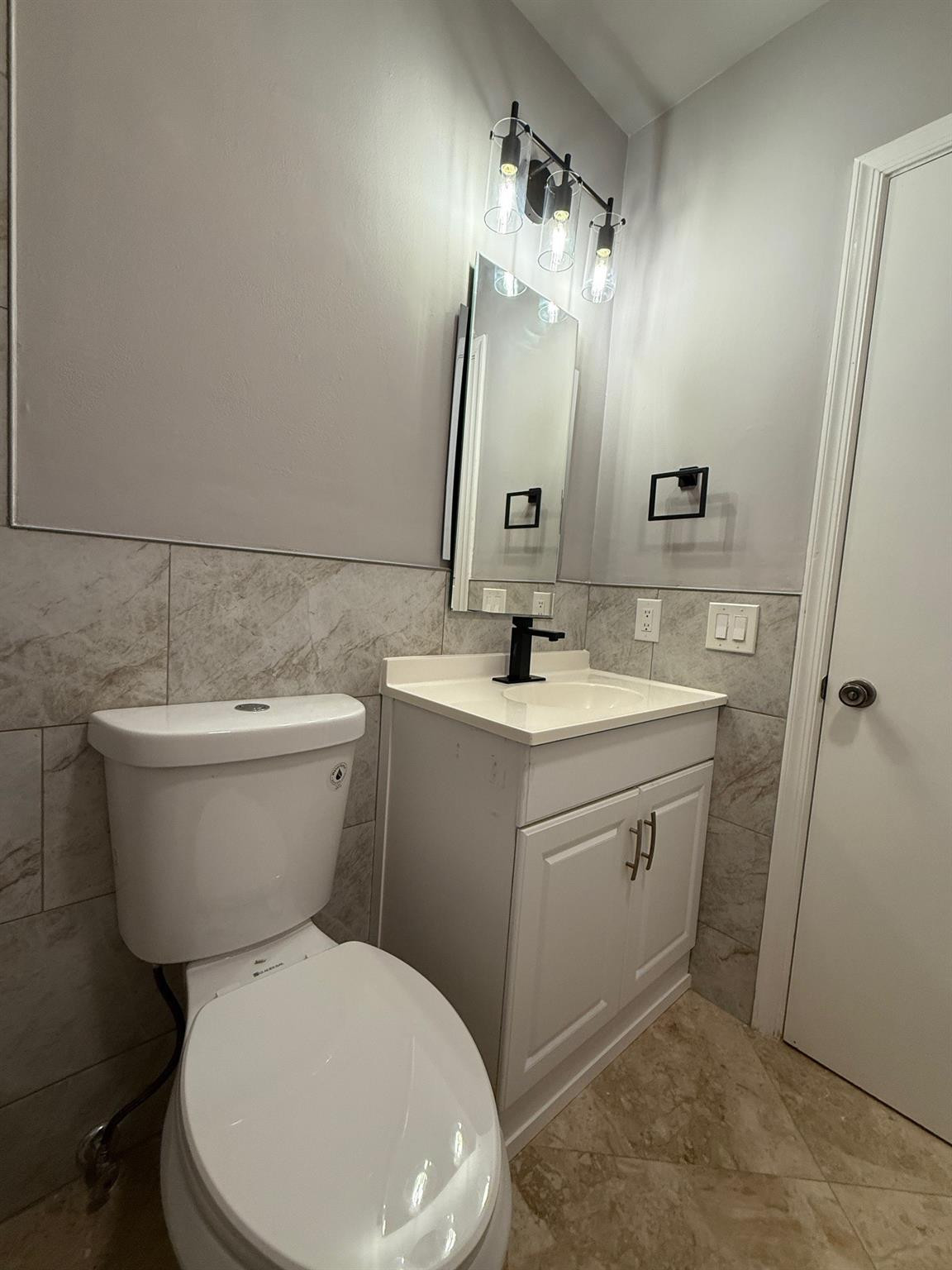Bathroom with white toilet and vanity, gray marble-look tile, and a three-light fixture above the mirror.