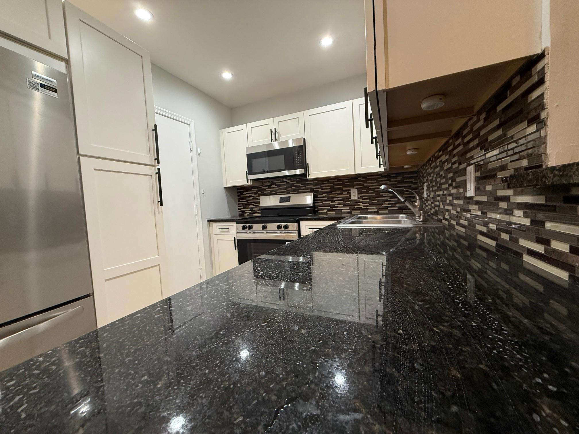 Kitchen with white cabinets, stainless appliances, dark mosaic backsplash, and glossy granite counters.