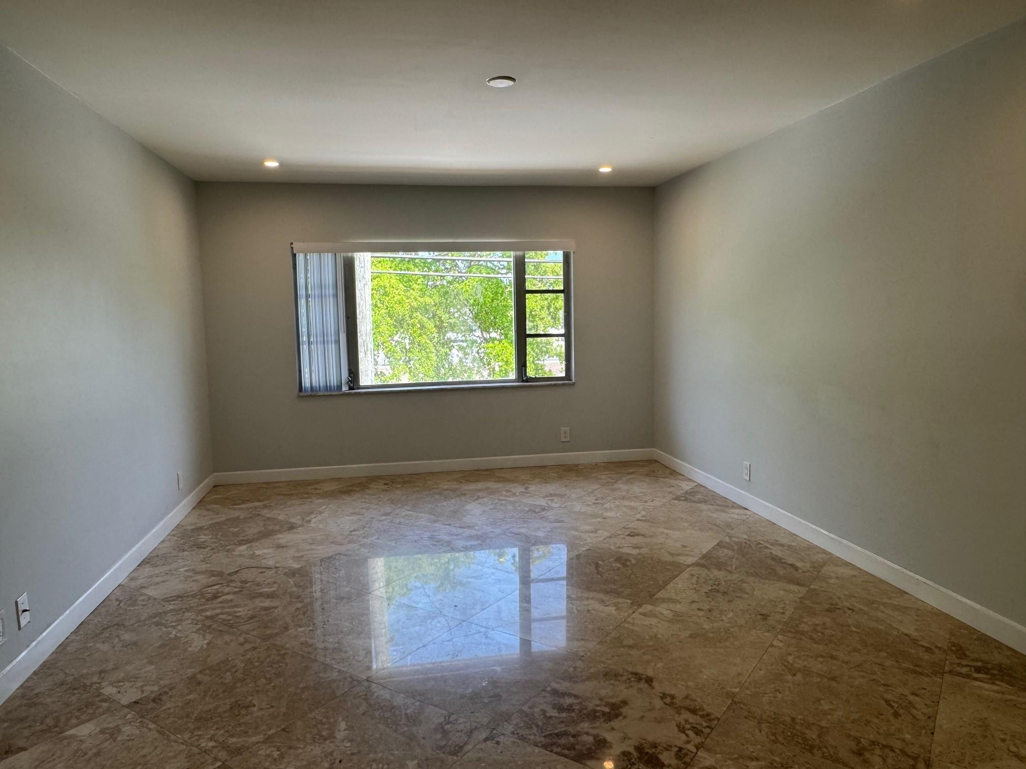 Empty room with glossy brown tile floors, recessed lights, and a window overlooking trees.