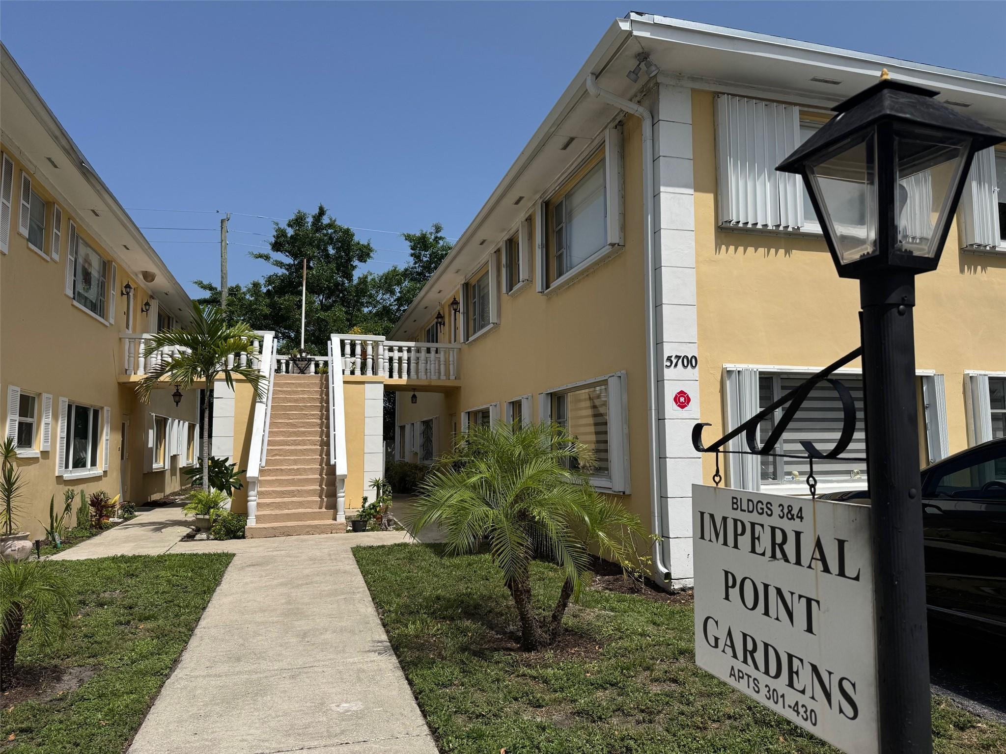 Yellow two-story apartment buildings flank a landscaped walkway and central staircase beside the Imperial Point Gardens sign.